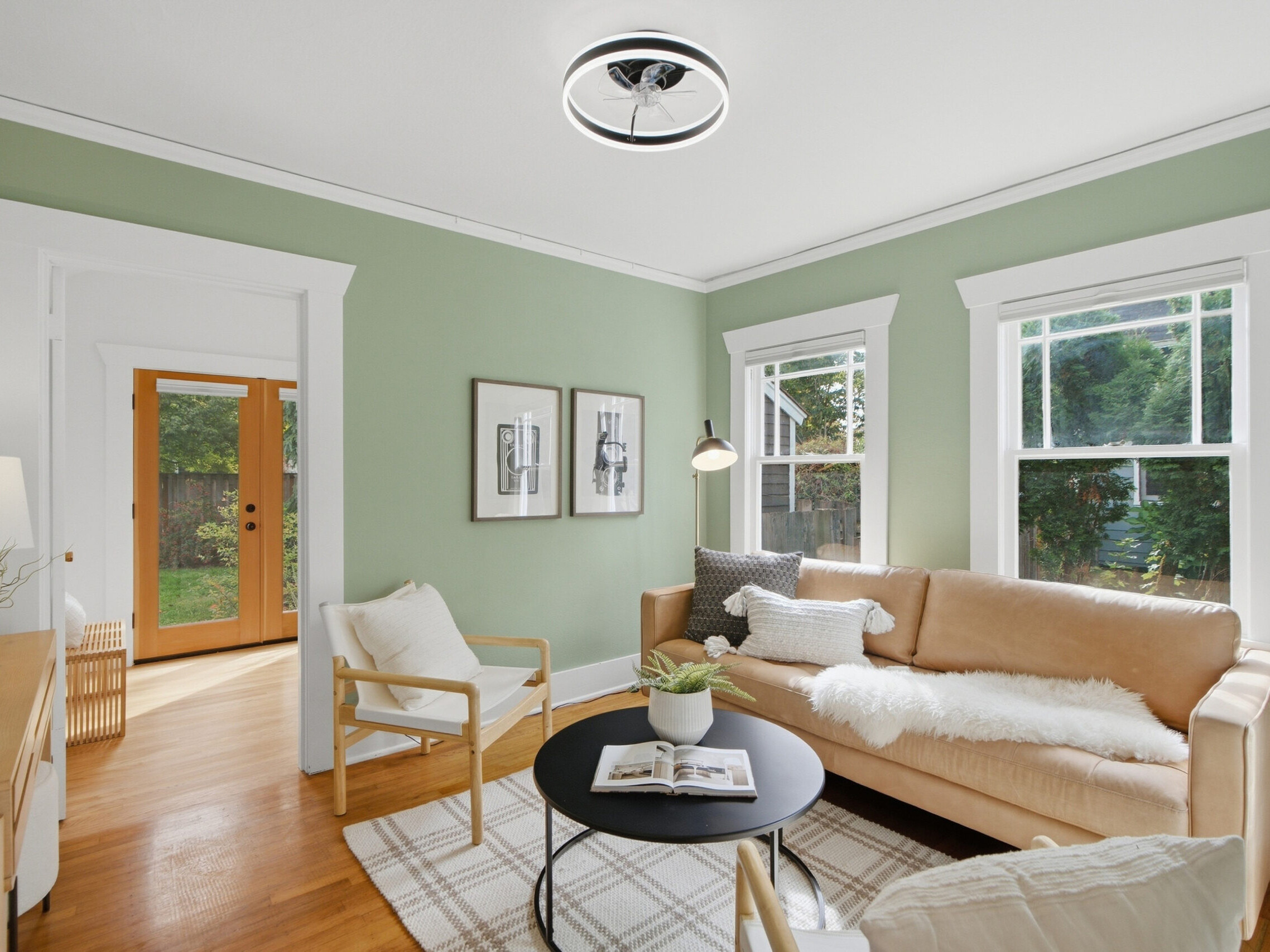 A cozy living room with light green walls, a beige sofa, two armchairs, a round black coffee table, a rug, and large windows letting in natural light. Two framed pictures hang on the wall near a door with glass panes.