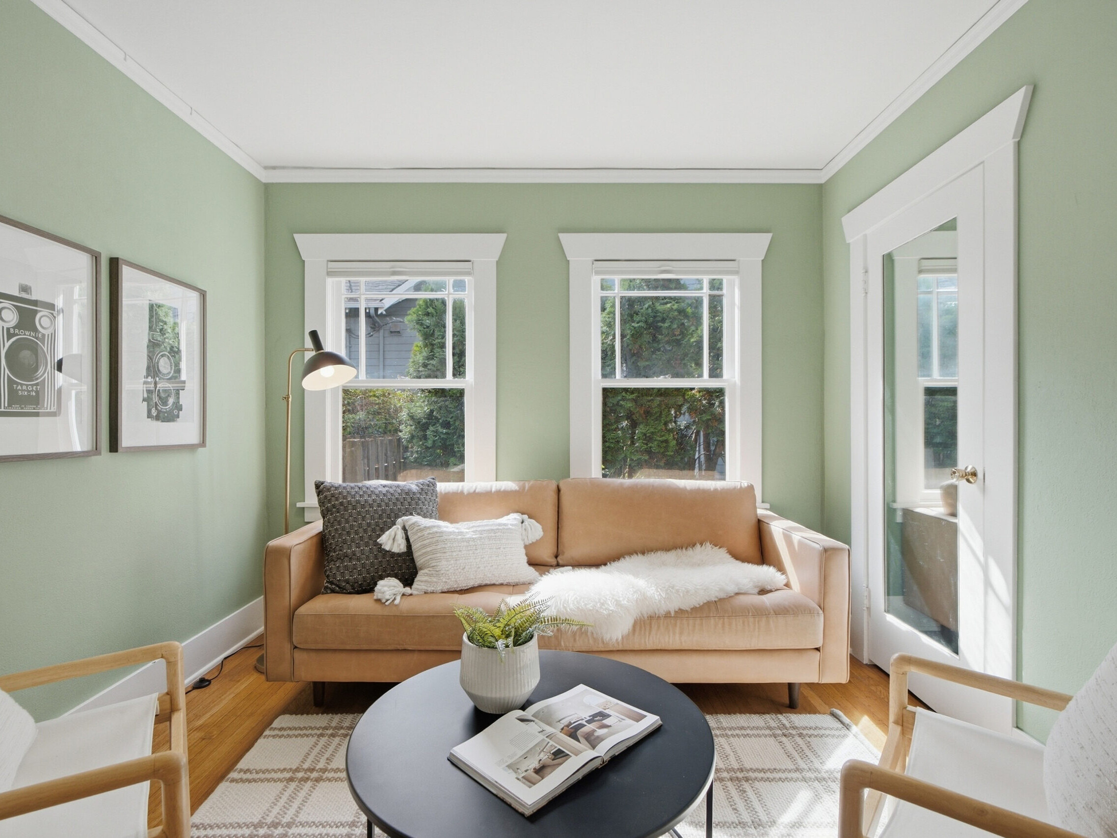 Bright living room with pale green walls, tan sofa, white fur throw, black round coffee table with books, white chairs, framed art, and large windows letting in natural light.