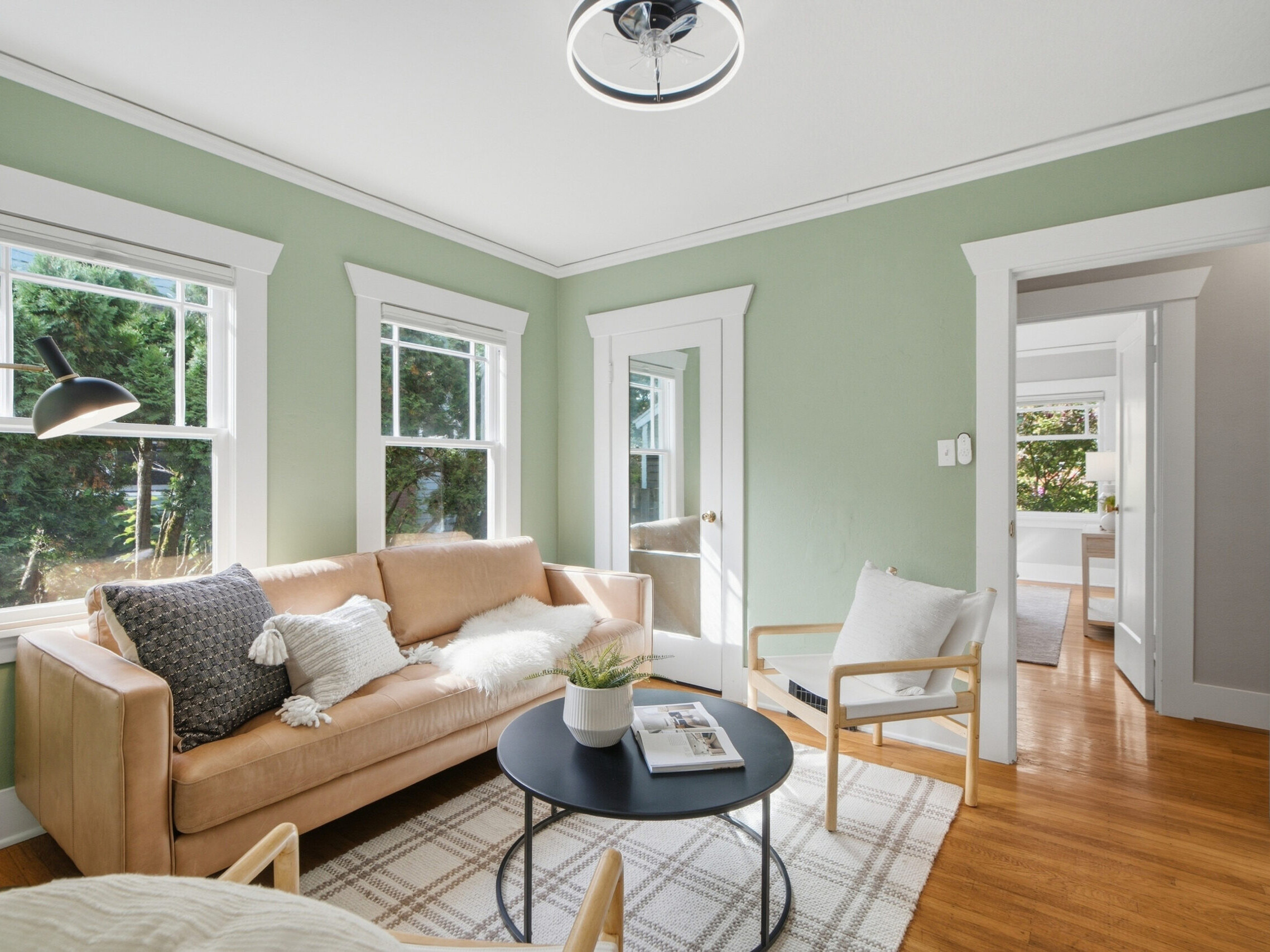 Bright living room with green walls, tan sofa, light wood chairs, a black round coffee table, white trim, large windows, and hardwood floors. A modern lamp and potted plant add decorative touches.