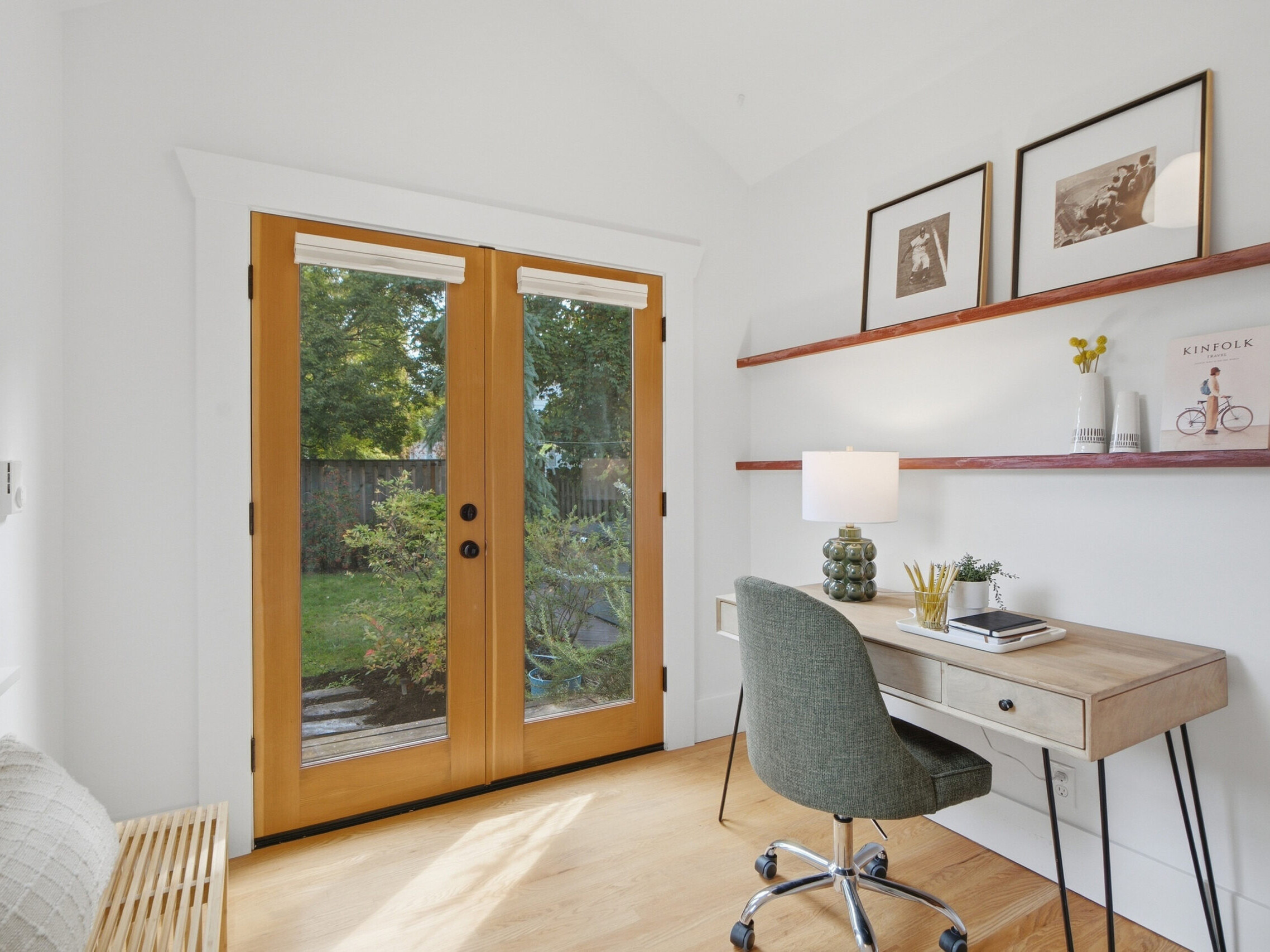 A bright home office with a wooden desk, green chair, shelves with decor, lamp, and framed art. Double glass doors open to a green backyard with trees.