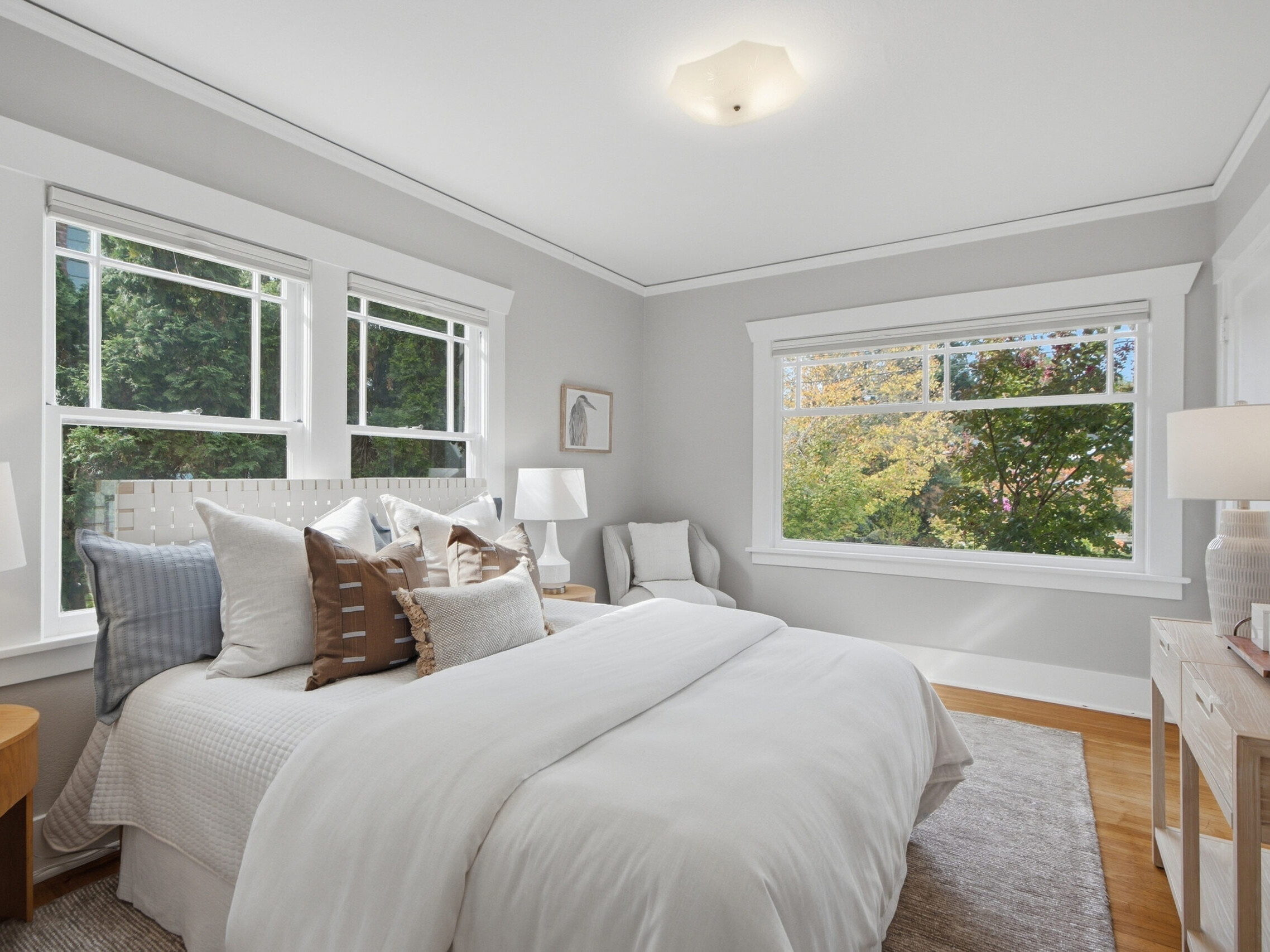 Bright bedroom with a large white bed, accent pillows, nightstands, lamps, and chairs by wide windows overlooking trees. Hardwood floors and light gray walls create a cozy, inviting atmosphere.