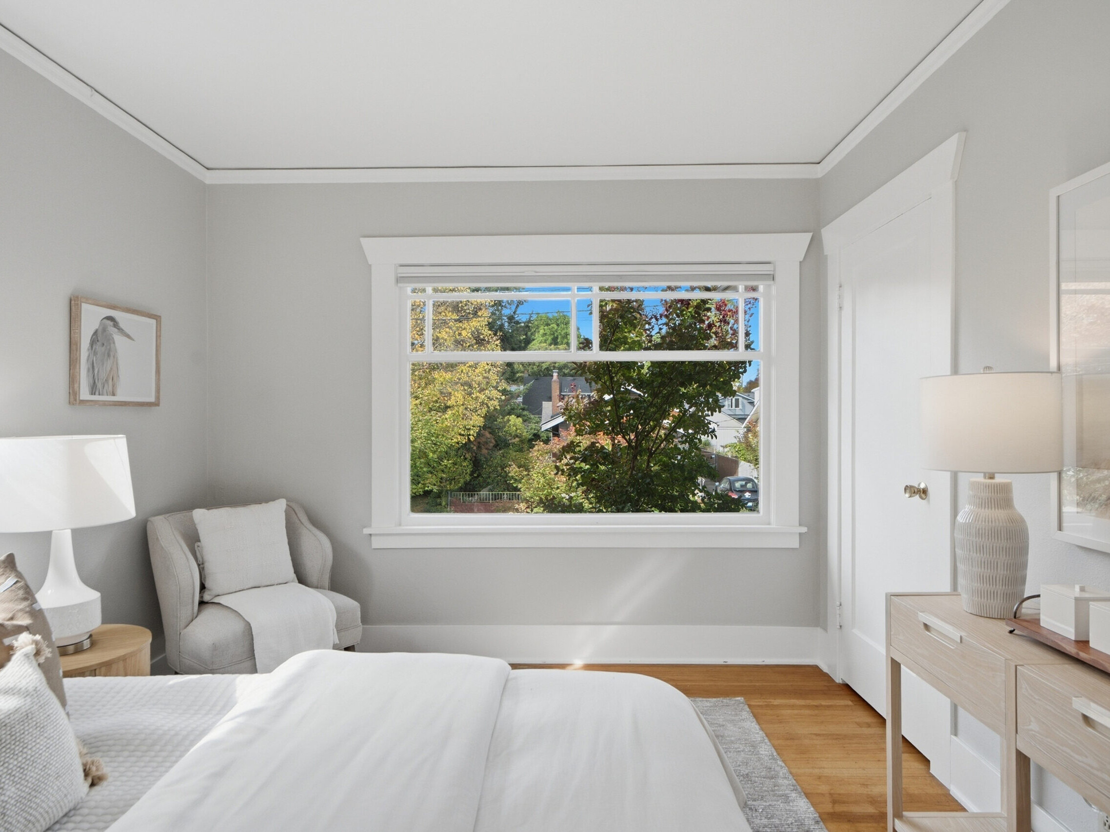 A bright bedroom with a large window showing green trees outside, a cozy armchair, bedside table with a lamp, and a dresser with decor on a wooden floor. Walls are light gray with white trim.