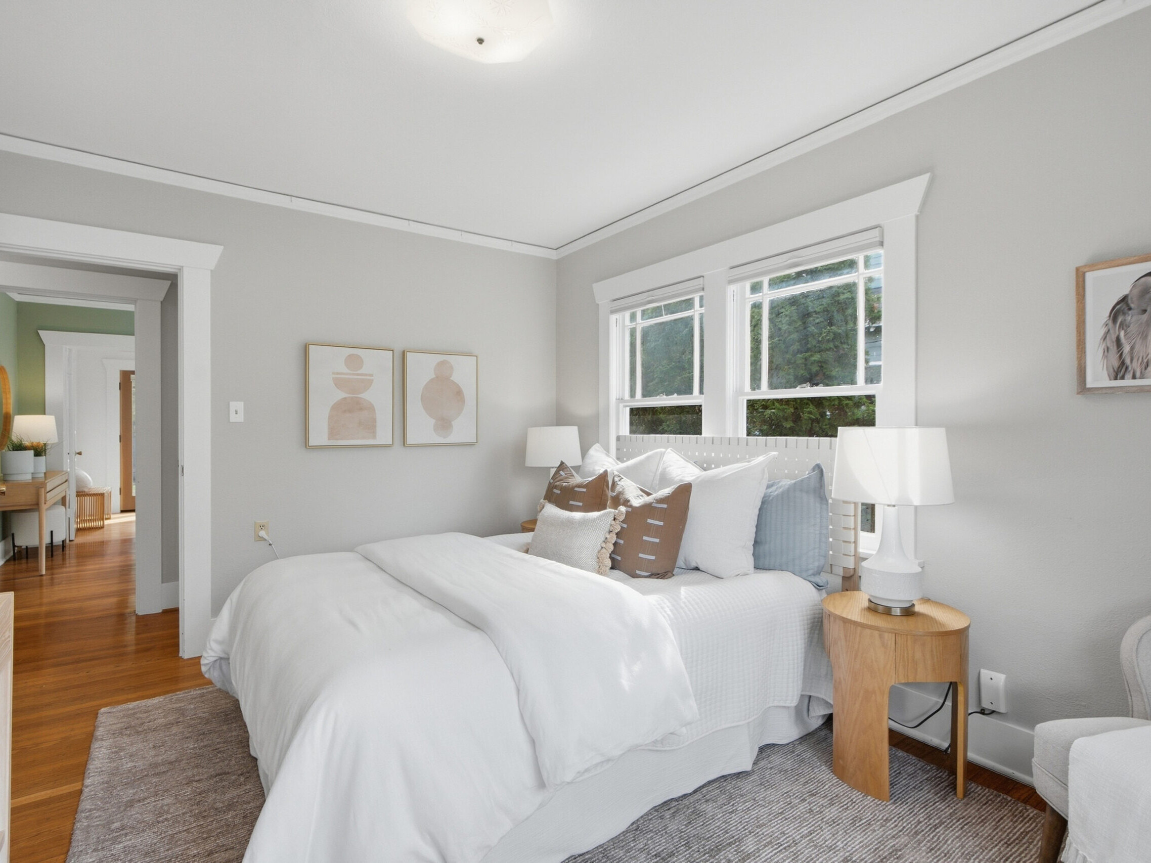 Bright bedroom with a neatly made bed, white bedding, decorative pillows, two nightstands with lamps, wall art, a gray armchair, and large windows letting in natural light.