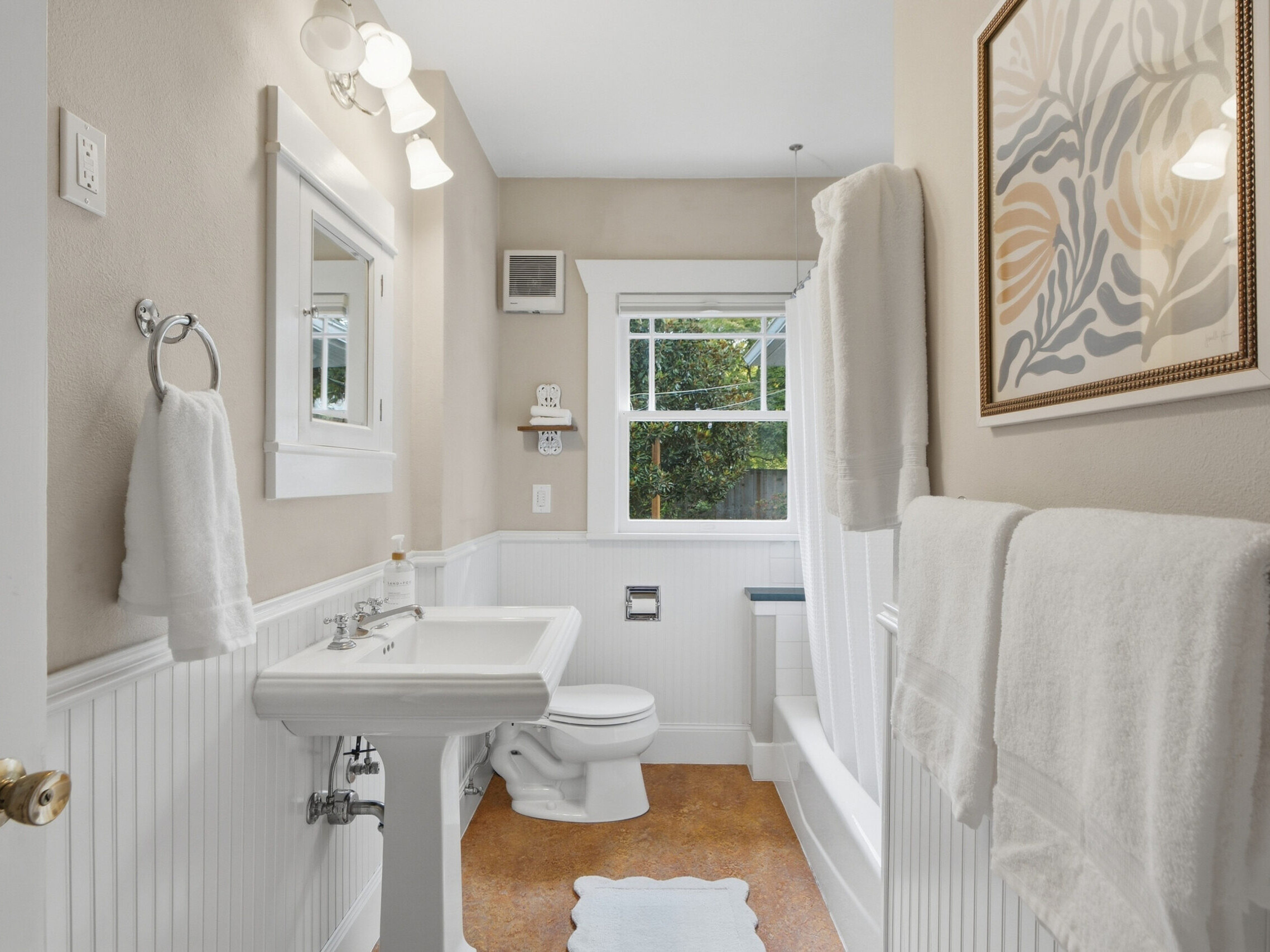 A bright bathroom with white fixtures, a pedestal sink, toilet, bathtub with a white shower curtain, towels, wall art, and a window letting in natural light.