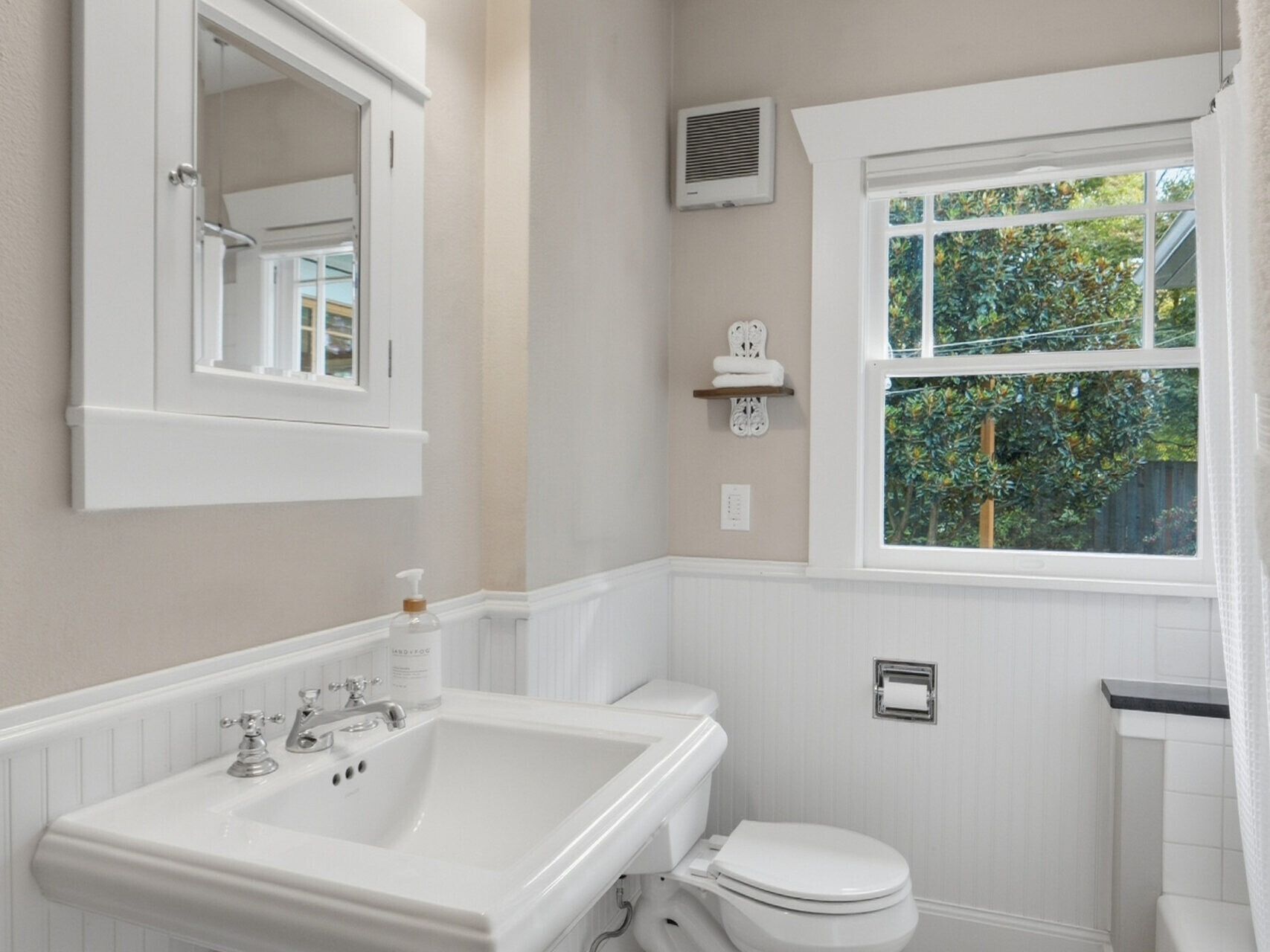 Bright bathroom with pedestal sink, white-framed mirror, toilet, towel ring, window with outdoor view, beige walls, white wainscoting, and brown floor. White bath mat lies on the floor near the sink.