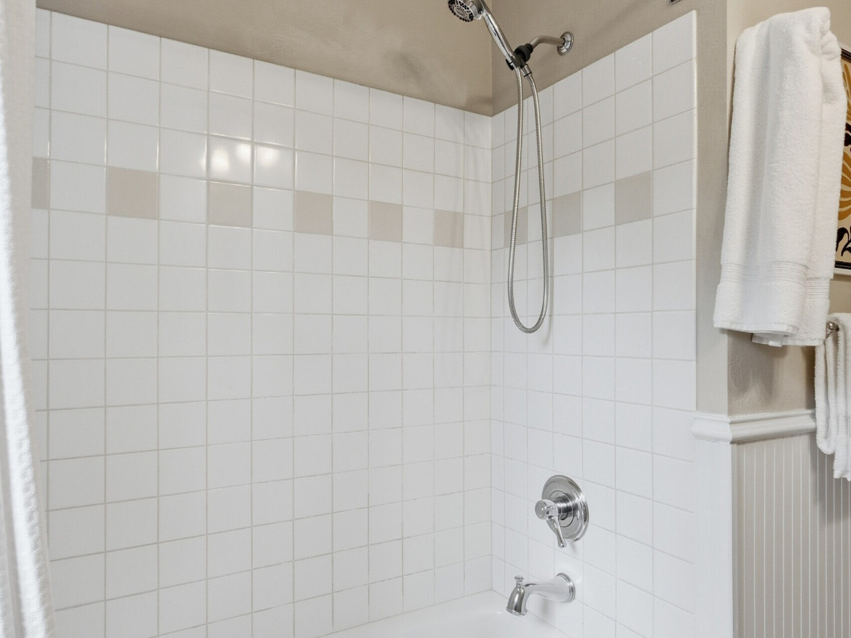 A white bathtub and shower combination with square white tiles, beige accent tiles, a silver showerhead, a white towel hanging, and beige walls.