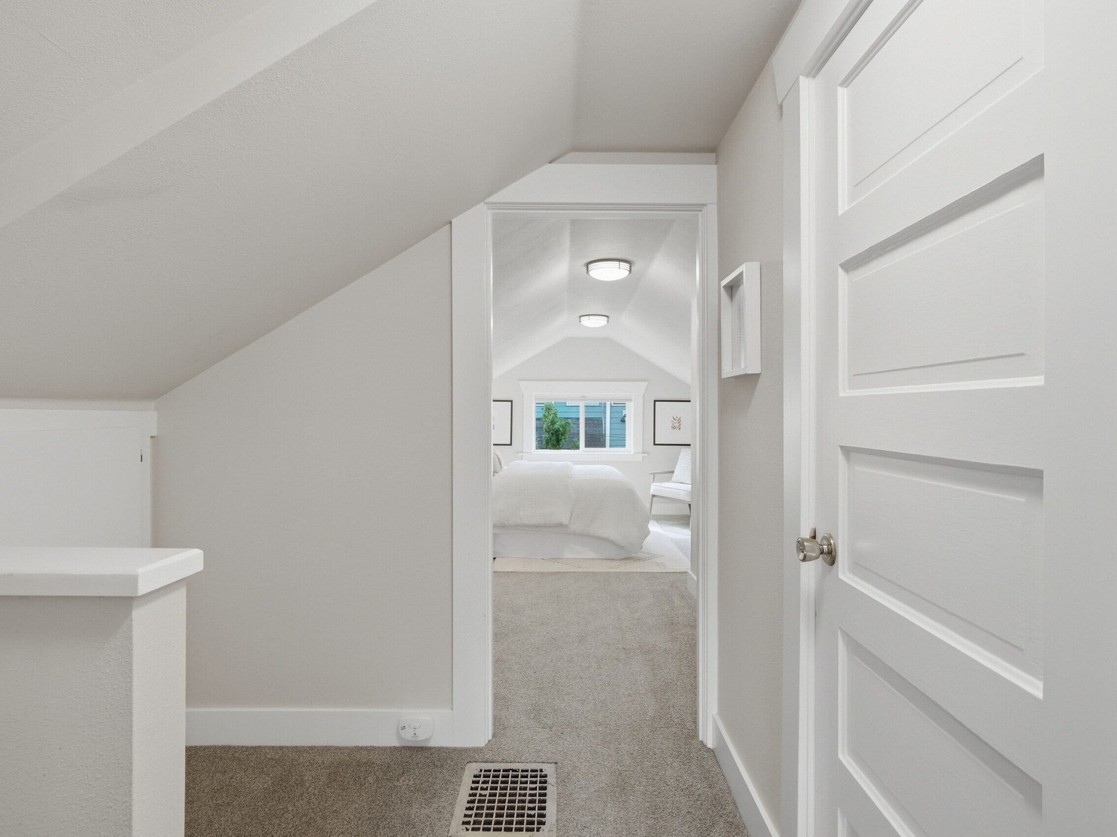 View from a hallway with sloped ceiling into a bright, carpeted bedroom with white walls, a bed, and large window letting in natural light. A white door is partially open in the foreground.