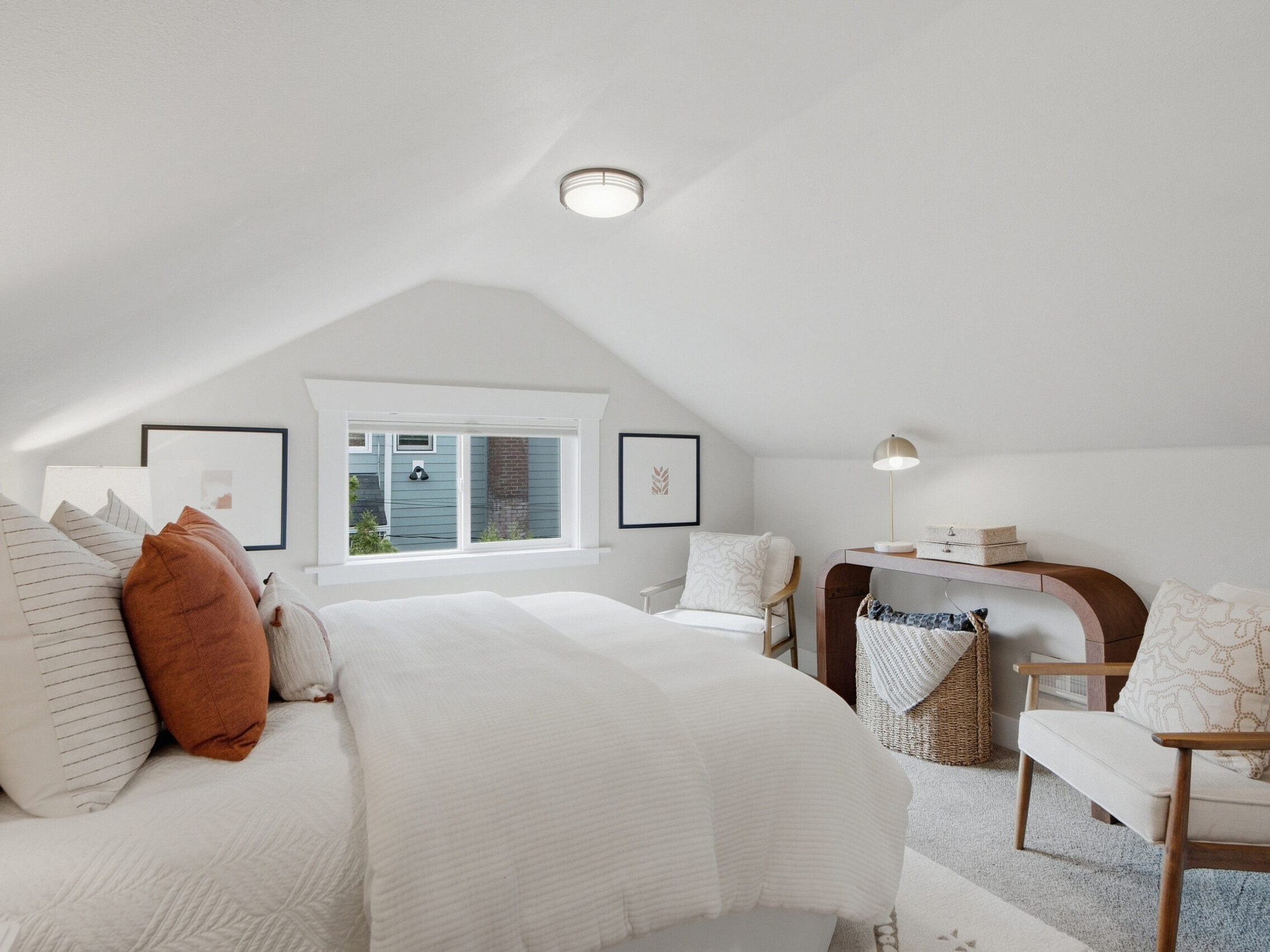 Bright attic bedroom with white walls, a large bed with white and rust pillows, two chairs, a small desk, woven baskets, and artwork. A window lets in natural light, creating an airy, cozy atmosphere.
