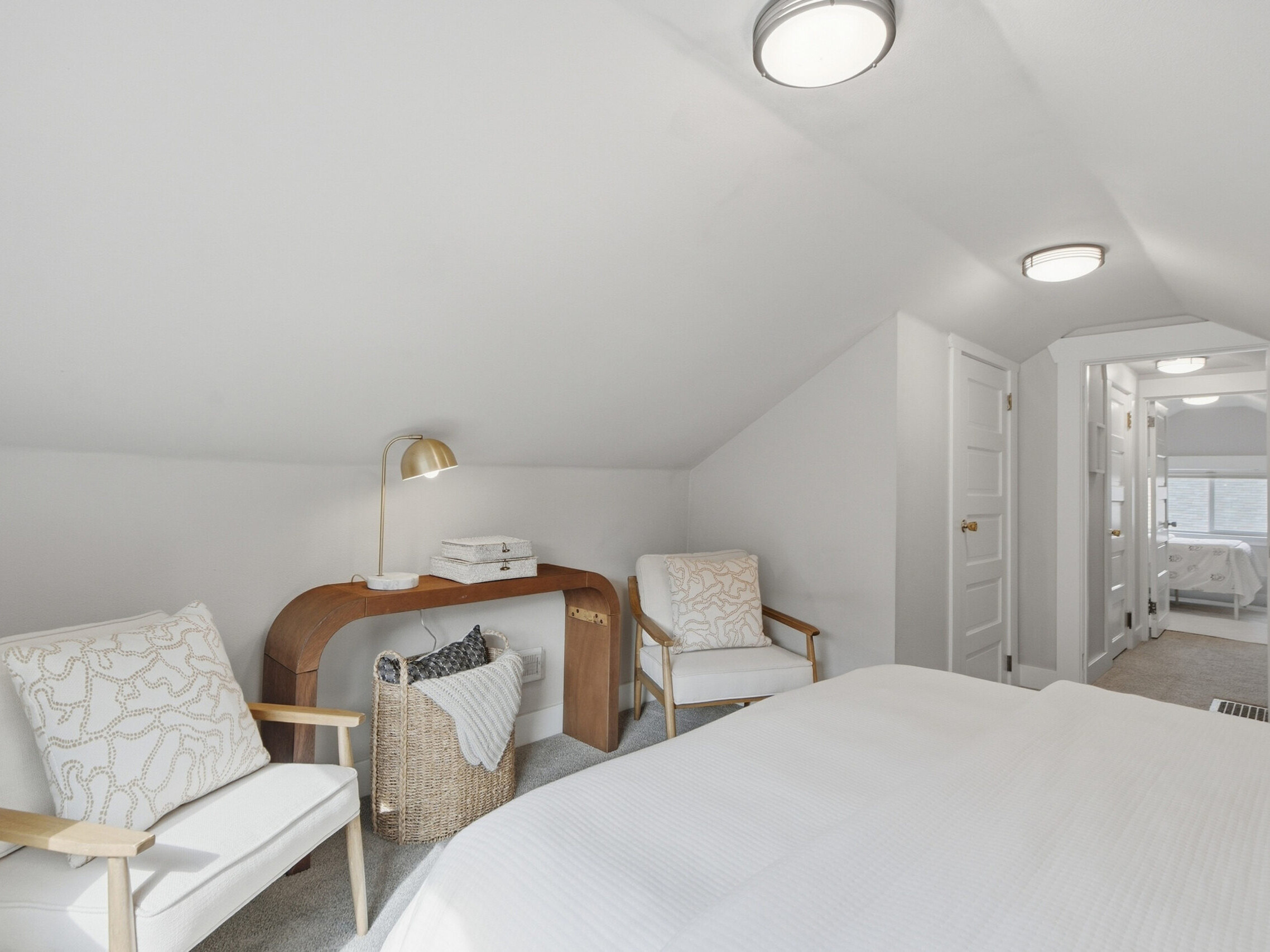 Bright attic bedroom with sloped ceiling, two white armchairs, a wooden desk with a lamp, woven basket, and a bed with white linens; hallway with doors and windows in the background.