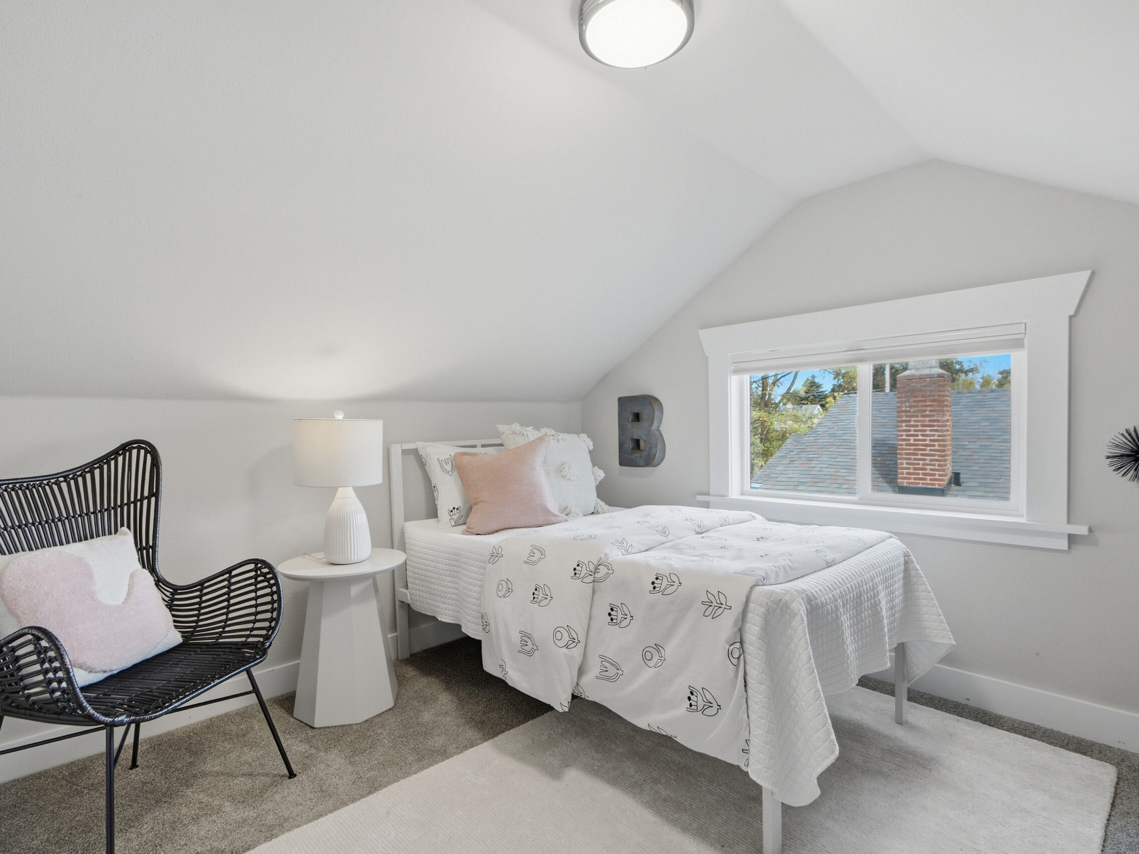 Bright, cozy bedroom with a twin bed, black wicker chair, white lamp on a round table, and a window showing a brick chimney. The room has white walls, gray carpet, and simple decor.