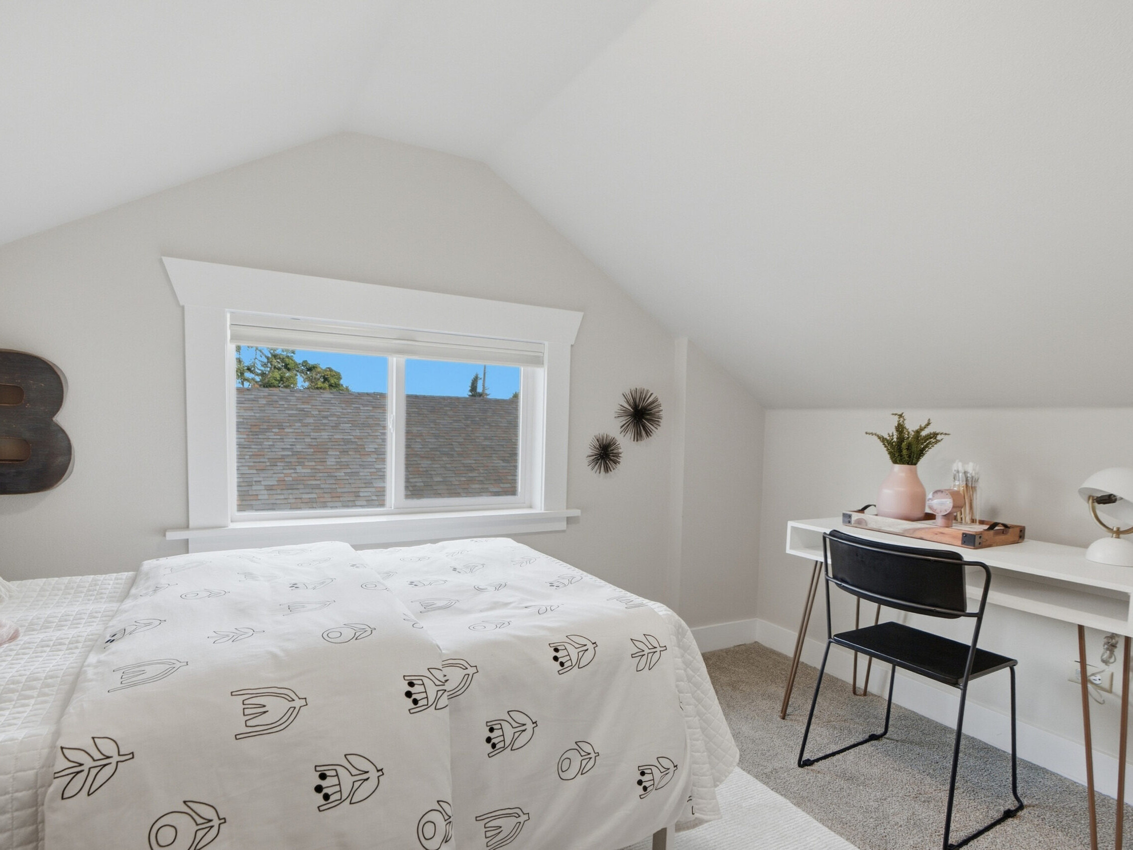 Bright bedroom with a bed, patterned white bedding, window, white desk with lamp and decor, black chair, and wall art. Light walls and carpet create an airy, cozy atmosphere.