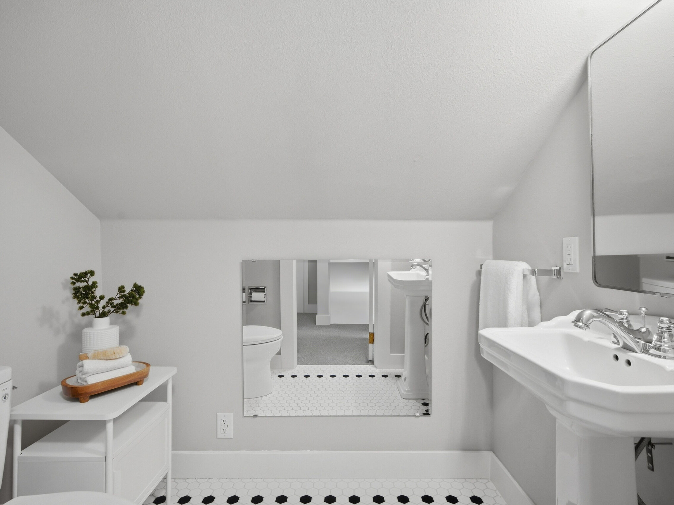Small, white bathroom with slanted ceiling, pedestal sink, wall mirror, towel rack, toilet, and a small shelf with rolled towels and a plant. The floor has a white tile pattern with black accents.