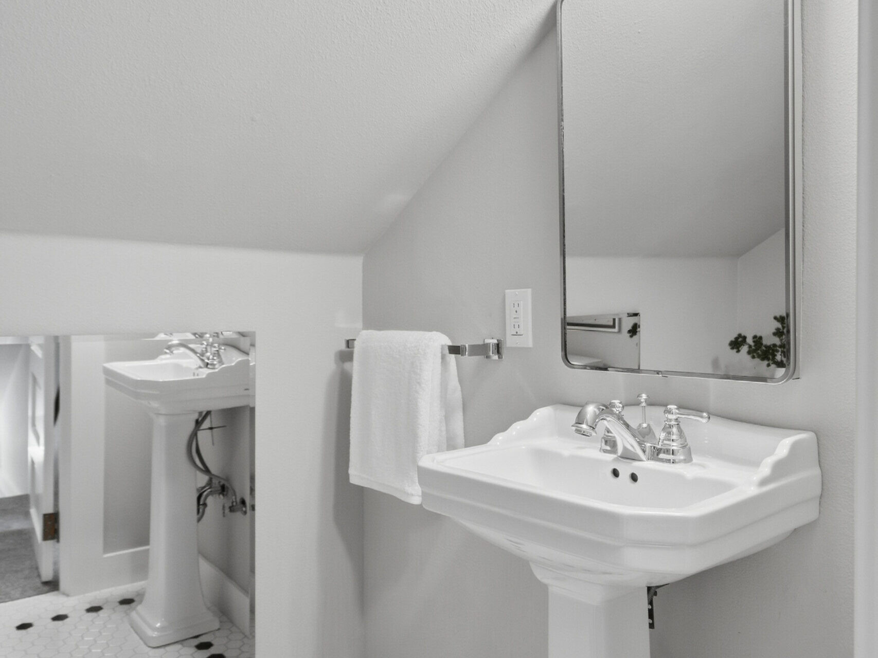 White pedestal sink with a rectangular mirror above it, small hexagonal black and white tile floor, a white towel on a metal rack, and a wall sconce light in a modern bathroom with angled ceiling.