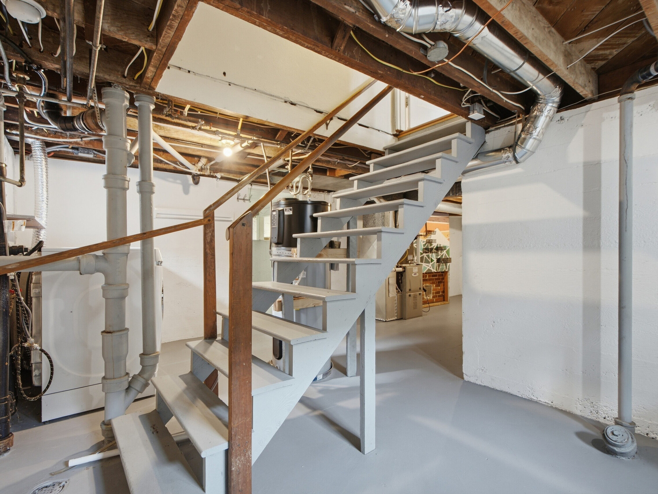 A clean, unfinished basement with exposed pipes and ductwork, gray painted stairs, white walls, and a concrete floor. Utilities and storage items are visible in the background.