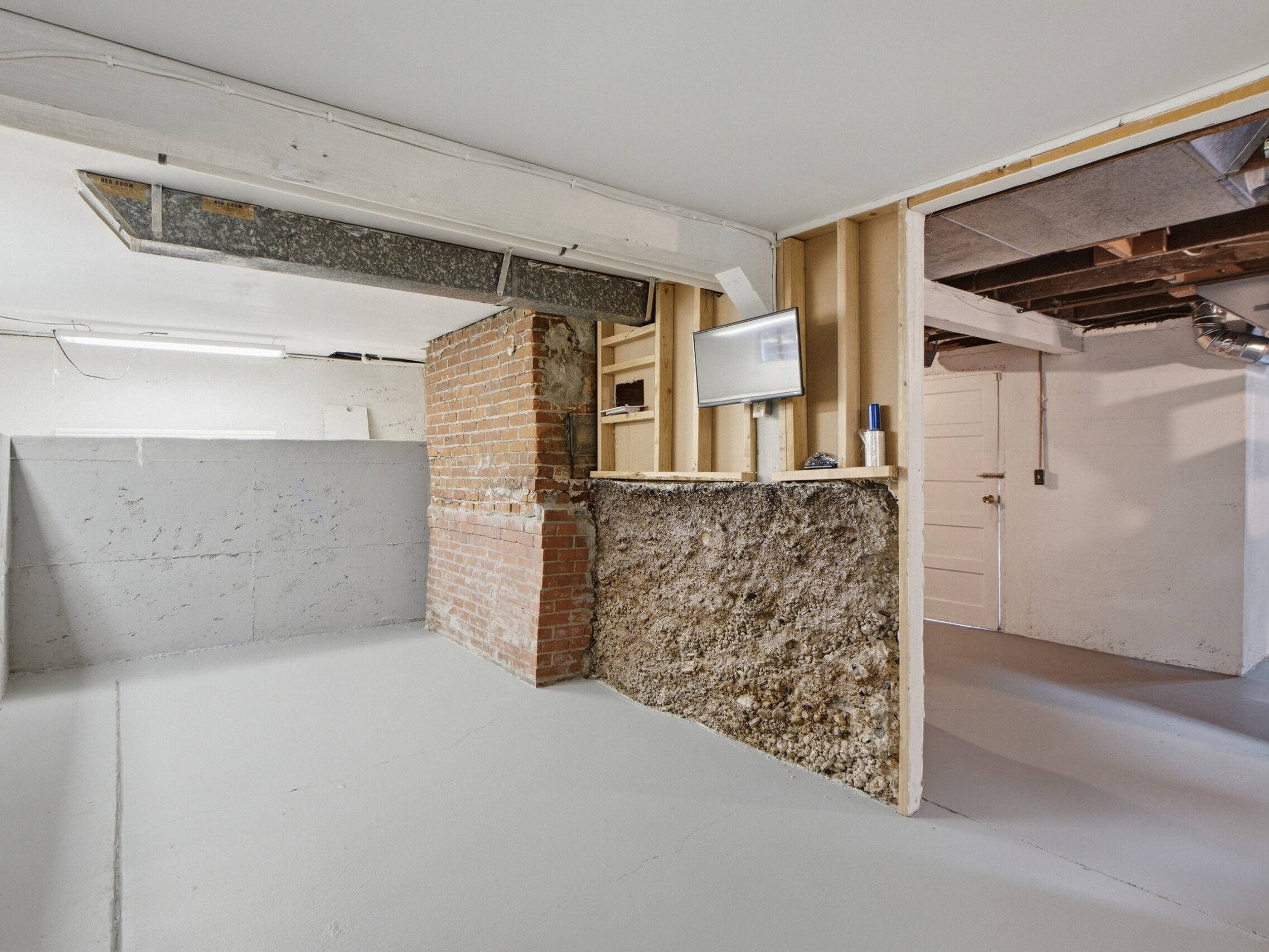 Unfinished basement with exposed brick, concrete walls, and open framing. Small mounted TV on the wall, white door in the back, and gray painted floor. Ceiling beams and ducts are visible.