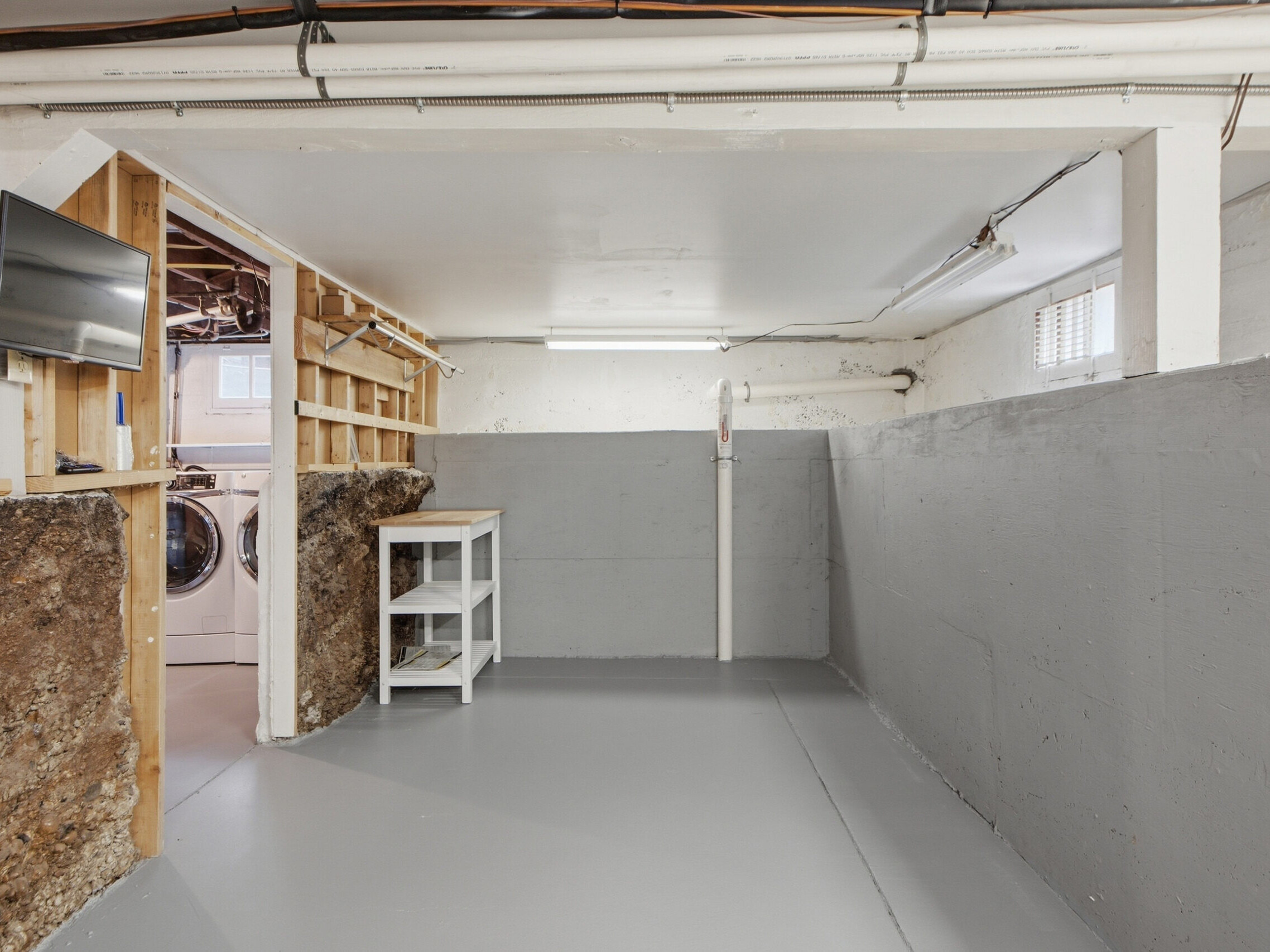 A clean, unfinished basement with gray painted floors and walls, exposed pipes, a wall-mounted TV, shelving, and a view into a laundry area with a washer and dryer. Light comes from ceiling fixtures and a small window.