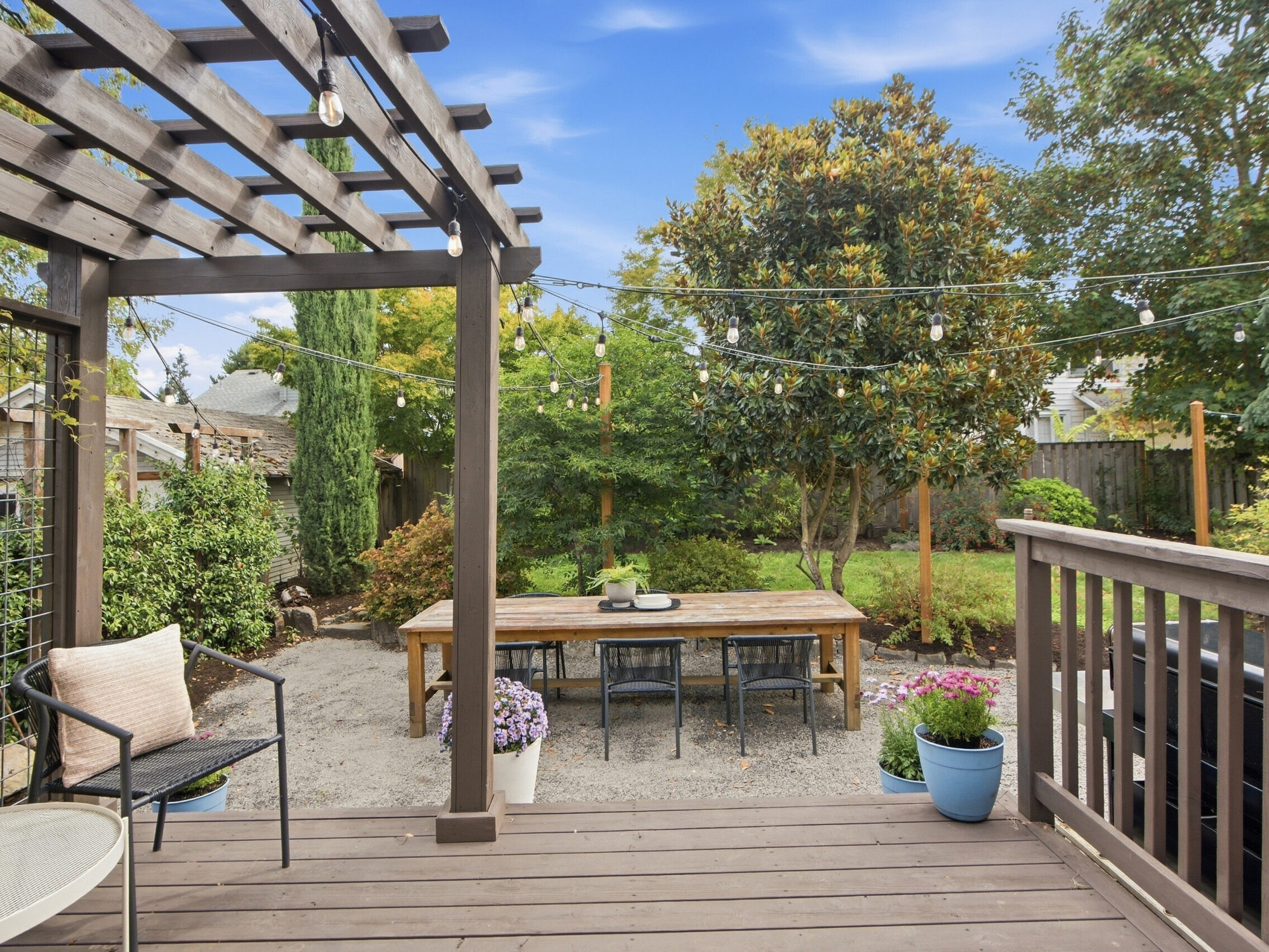 A wooden deck overlooks a backyard with a picnic table, string lights, potted plants, and lush green trees under a partly cloudy blue sky.