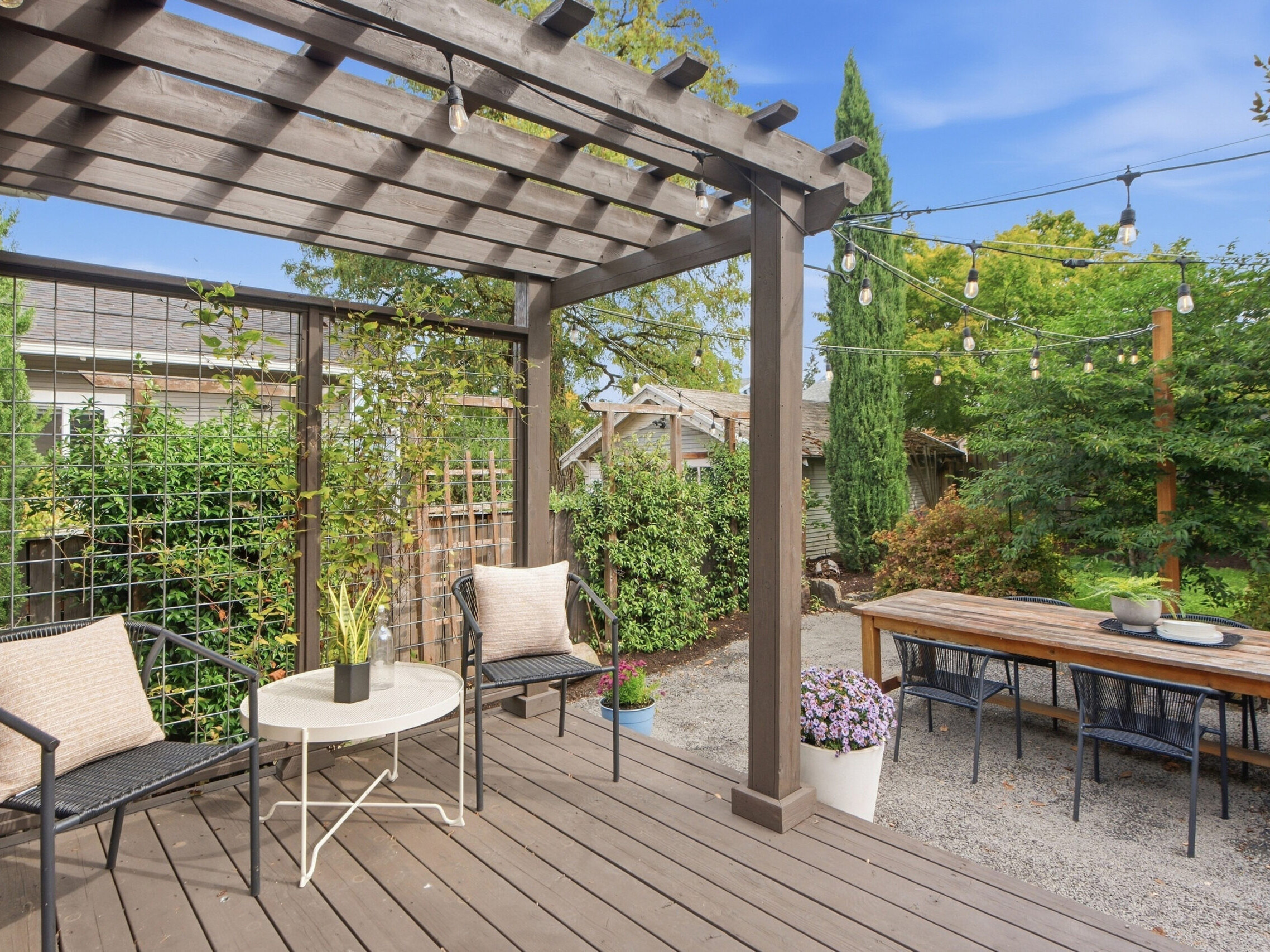 A wooden patio with a pergola, two chairs, and a small table, overlooking a garden with string lights, a long dining table, and various plants and trees.