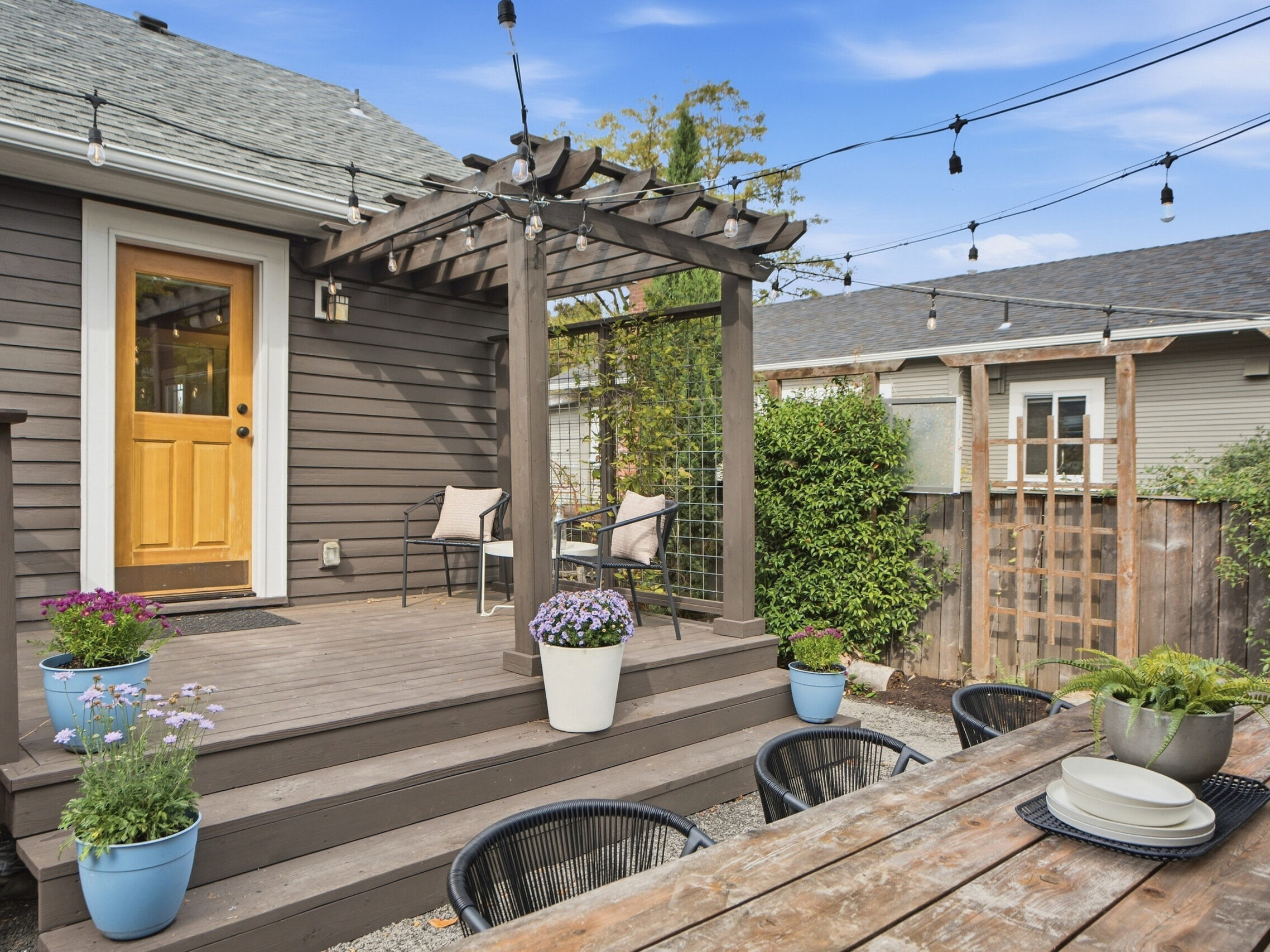 A cozy backyard patio with a pergola, string lights, potted plants, two chairs, and a wooden dining table set for four on a gravel area. The house has brown siding and a yellow door.