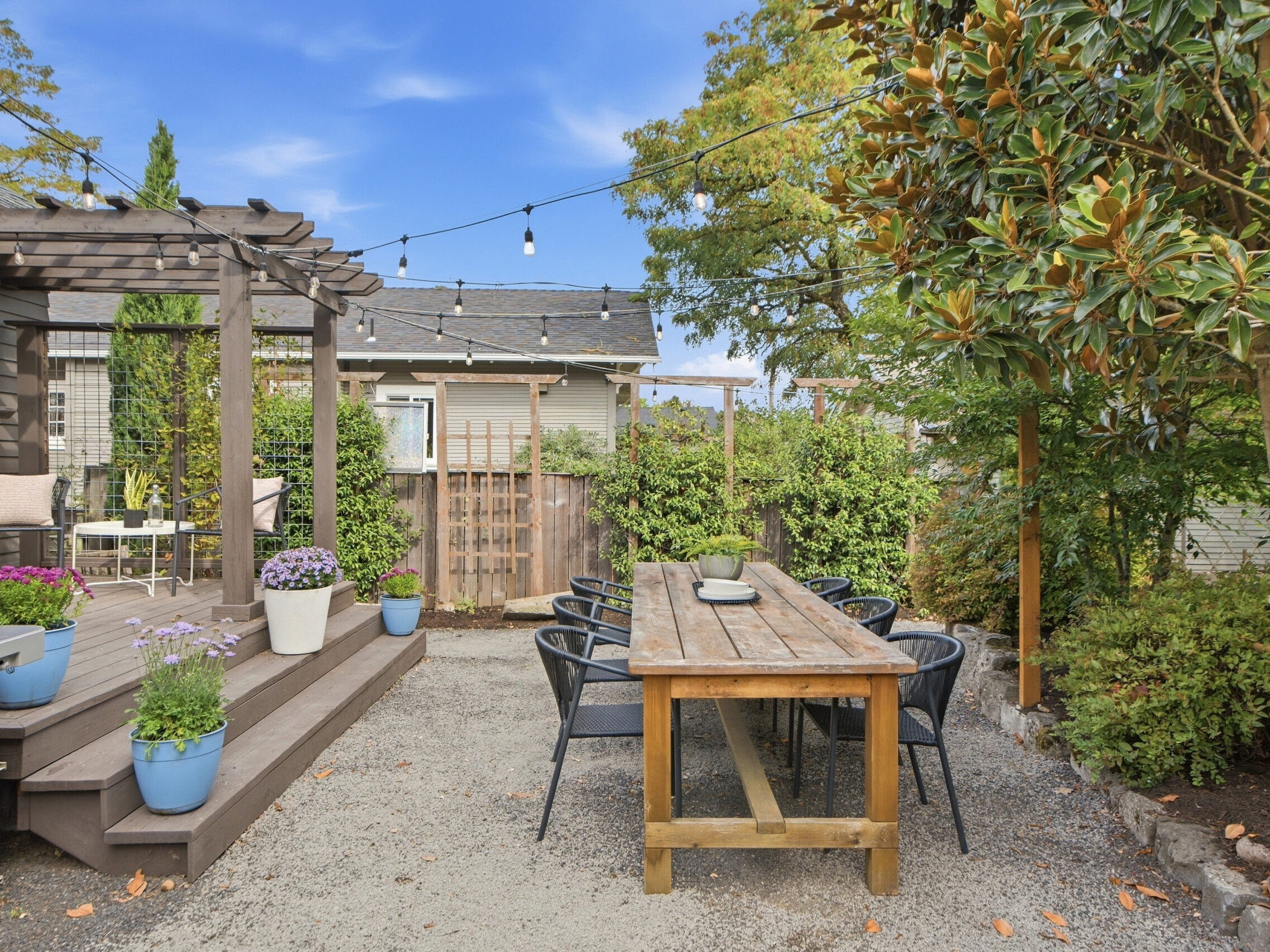 Outdoor patio with a wooden dining table, black chairs, string lights, potted plants, and a pergola. Lush greenery and a tree surround the cozy, inviting space under a blue sky.