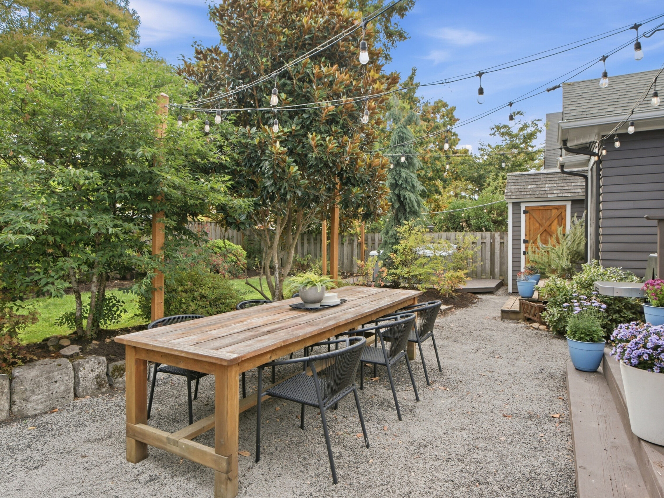 A rustic outdoor dining area with a long wooden table, black chairs, string lights overhead, potted plants, and greenery surrounding the space on a gravel patio.
