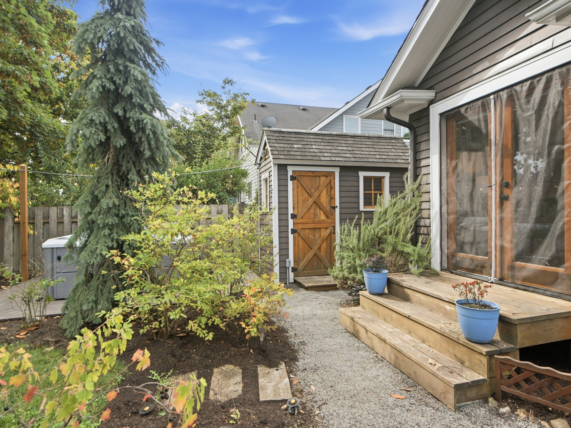 Wooden steps with potted plants lead to a house with brown siding and glass doors. A garden with shrubs and a tree surrounds a gravel path to a shed with a wooden door and fenced yard under a blue sky.