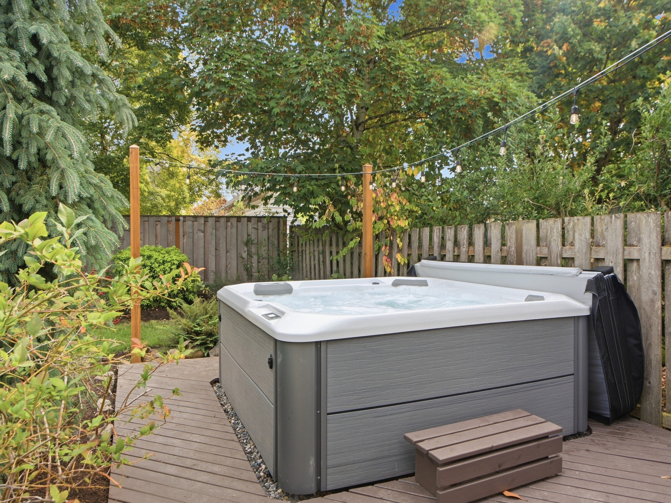 A gray hot tub filled with water sits on a wooden deck in a fenced backyard, surrounded by green trees and plants under a partly cloudy sky. A small set of steps leads to the hot tub.