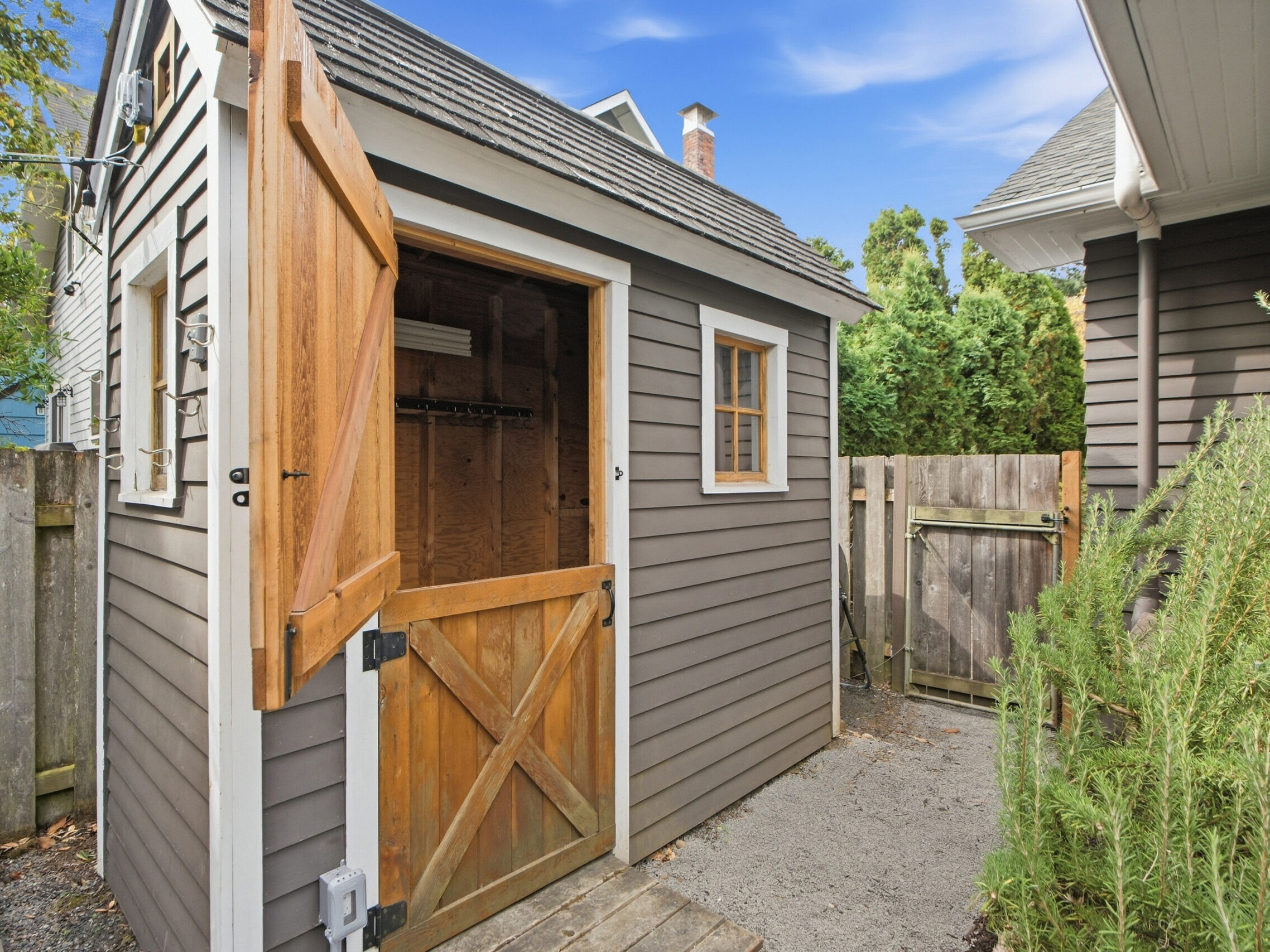 Small gray shed with a wooden Dutch door partially open, revealing a simple interior. Theres a wooden fence, some greenery, and part of a house visible nearby under a blue sky.