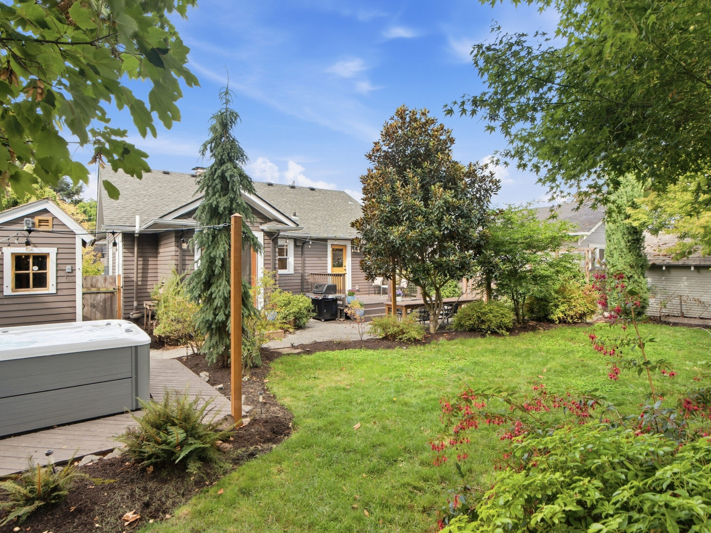 Backyard with green lawn, trees, shrubs, a wooden deck with a hot tub, and a brown house with a gable roof under a blue sky with scattered clouds.