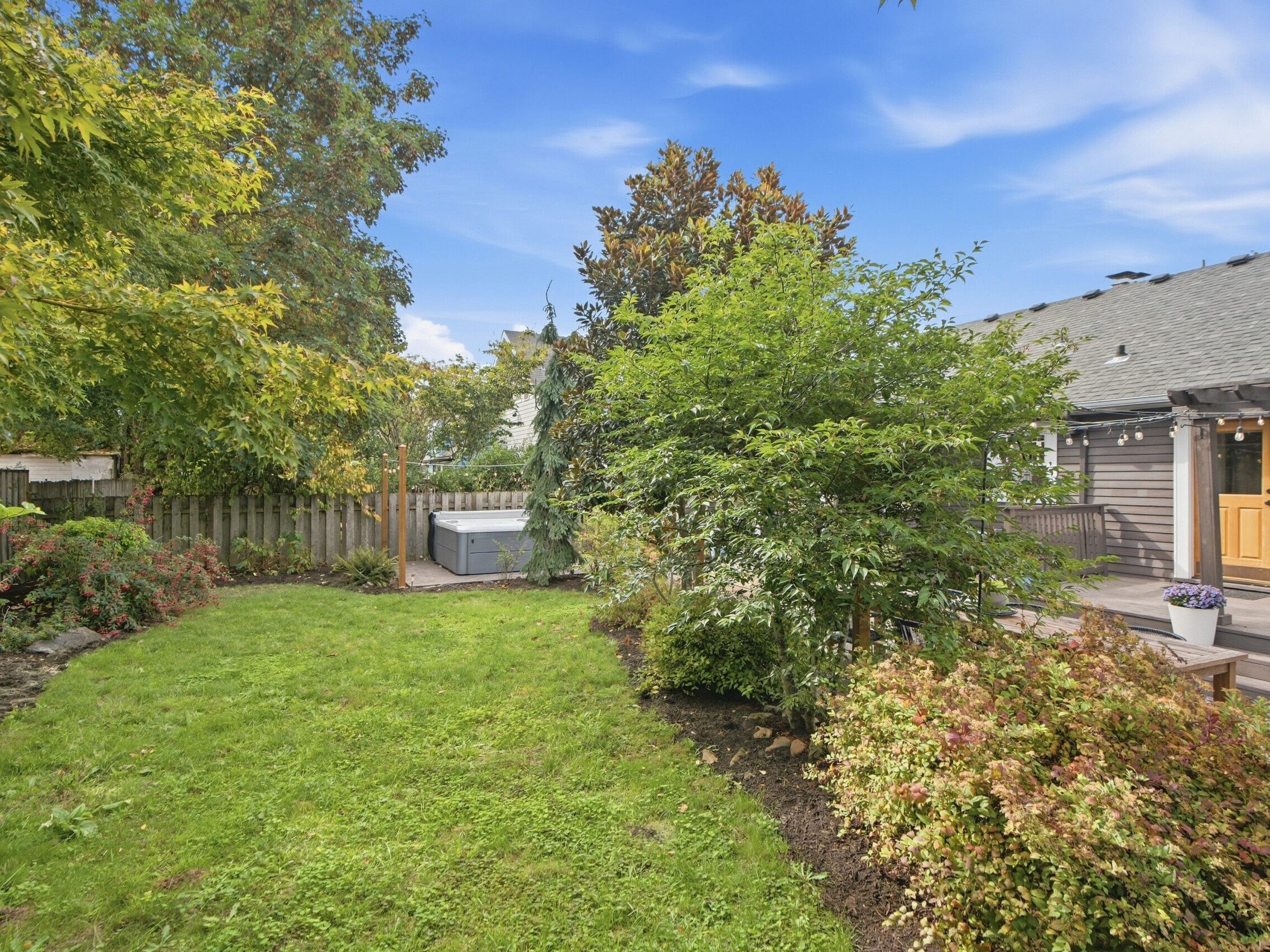 A backyard with green grass, shrubs, and trees, bordered by a wooden fence. Theres a hot tub in the corner and a wooden deck attached to a house with a yellow door. The sky is blue with some clouds.