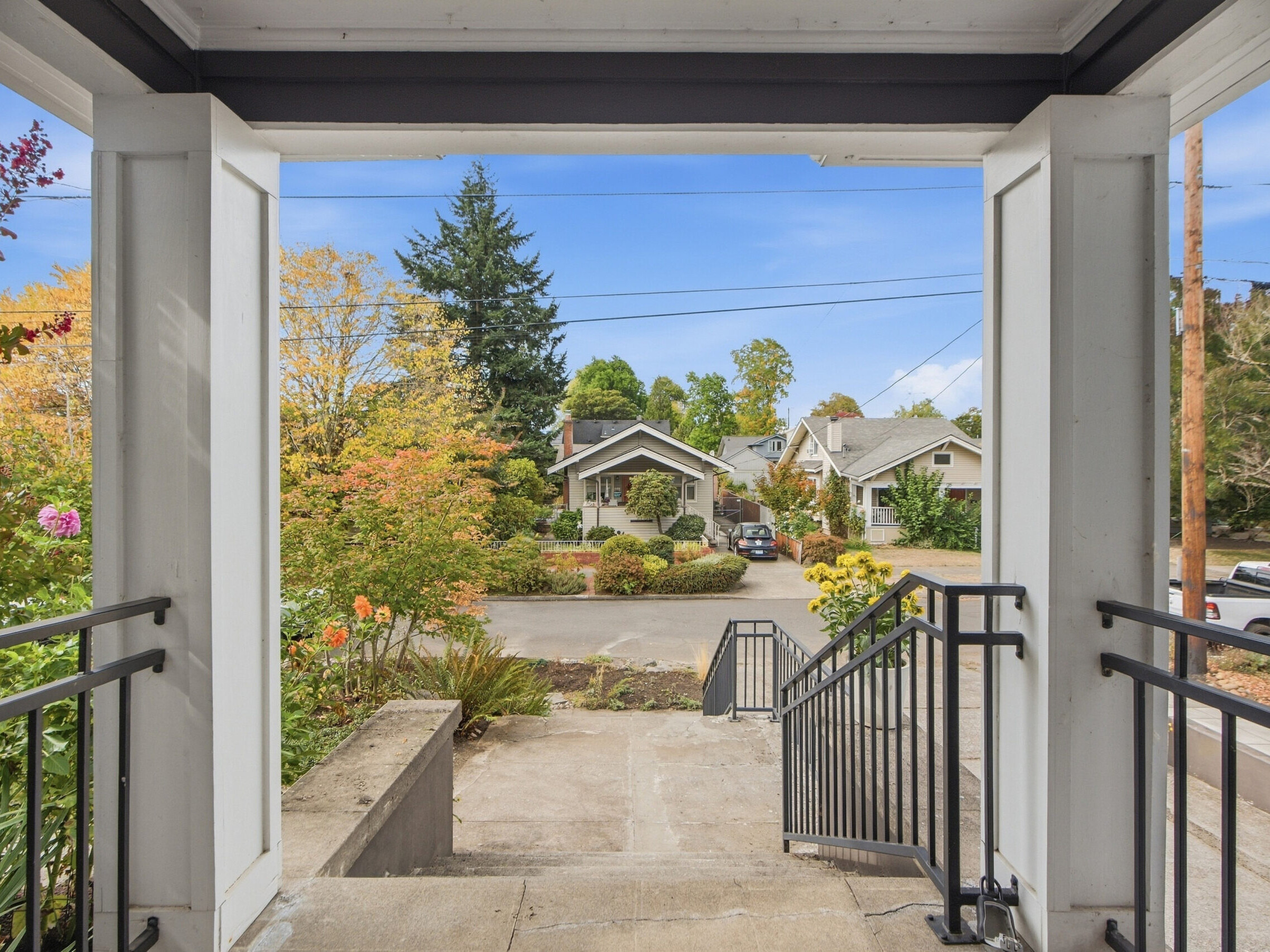 View from a porch looking out onto a residential street lined with houses, trees with autumn foliage, and a blue sky. Black railings line the porch steps.