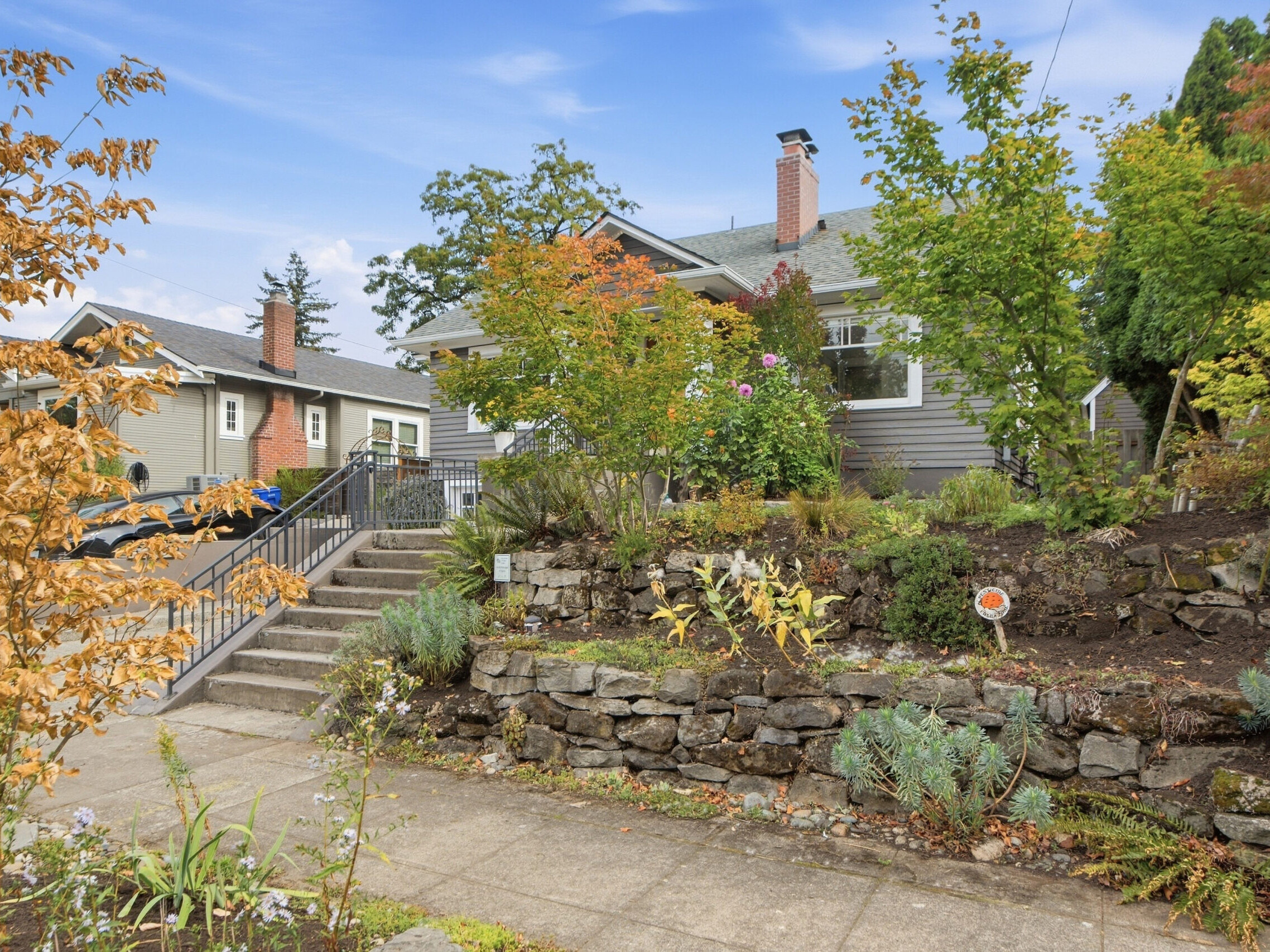 Gray house with a sloped front yard, stone retaining wall, stairway, and various trees and plants. A sidewalk and neighboring house are visible under a blue sky with scattered clouds.