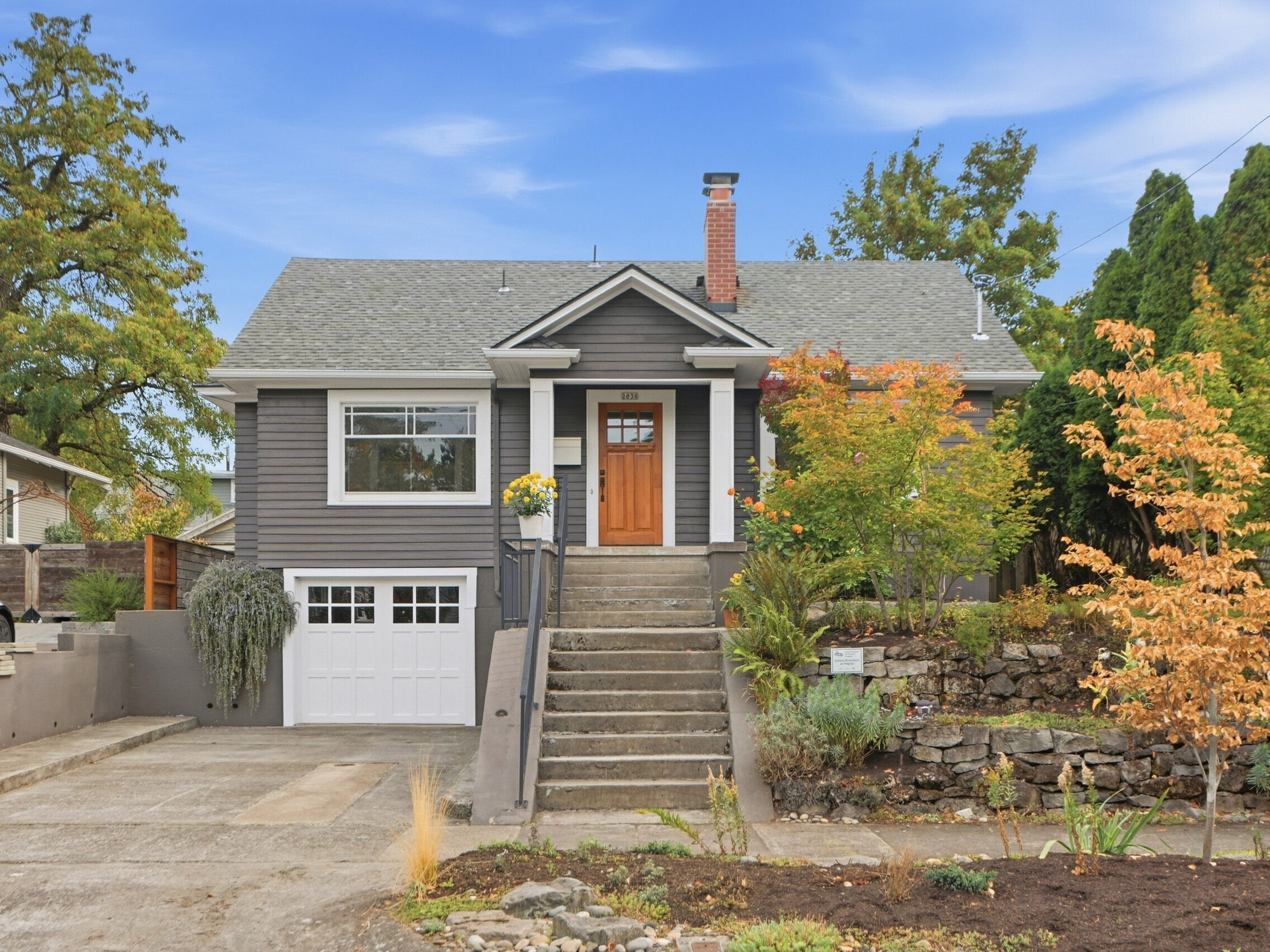 Gray house with white trim, a front porch, and a red door. There are steps leading up to the entry, a single-car garage, and landscaped front yard with trees and shrubs. Blue sky and neighboring homes are visible.