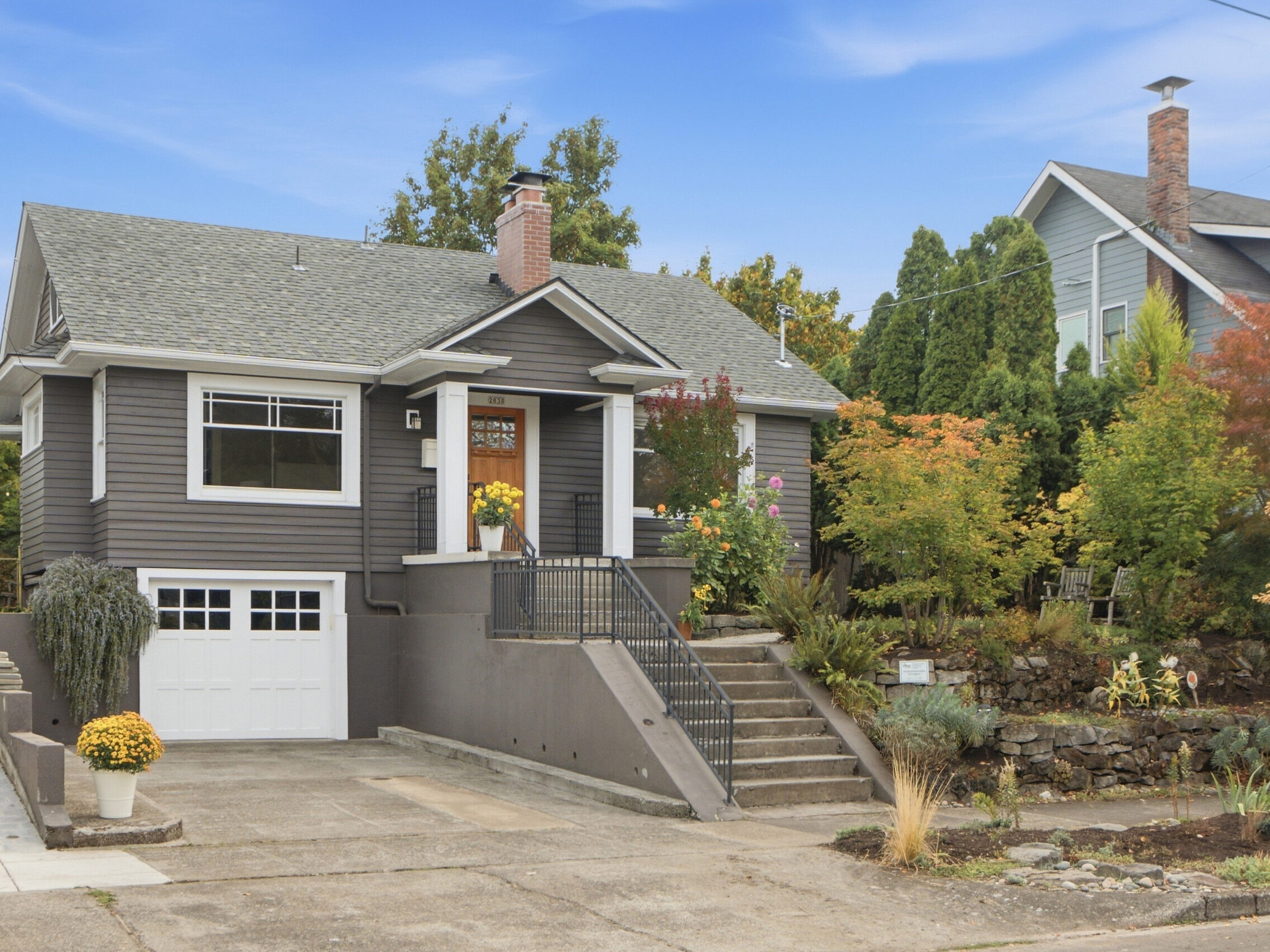Gray house with white trim, a front porch, and stairs leading up from the sidewalk. There is a garage, driveway, and colorful landscaping with autumn foliage and flowers. Another house is visible on the right.