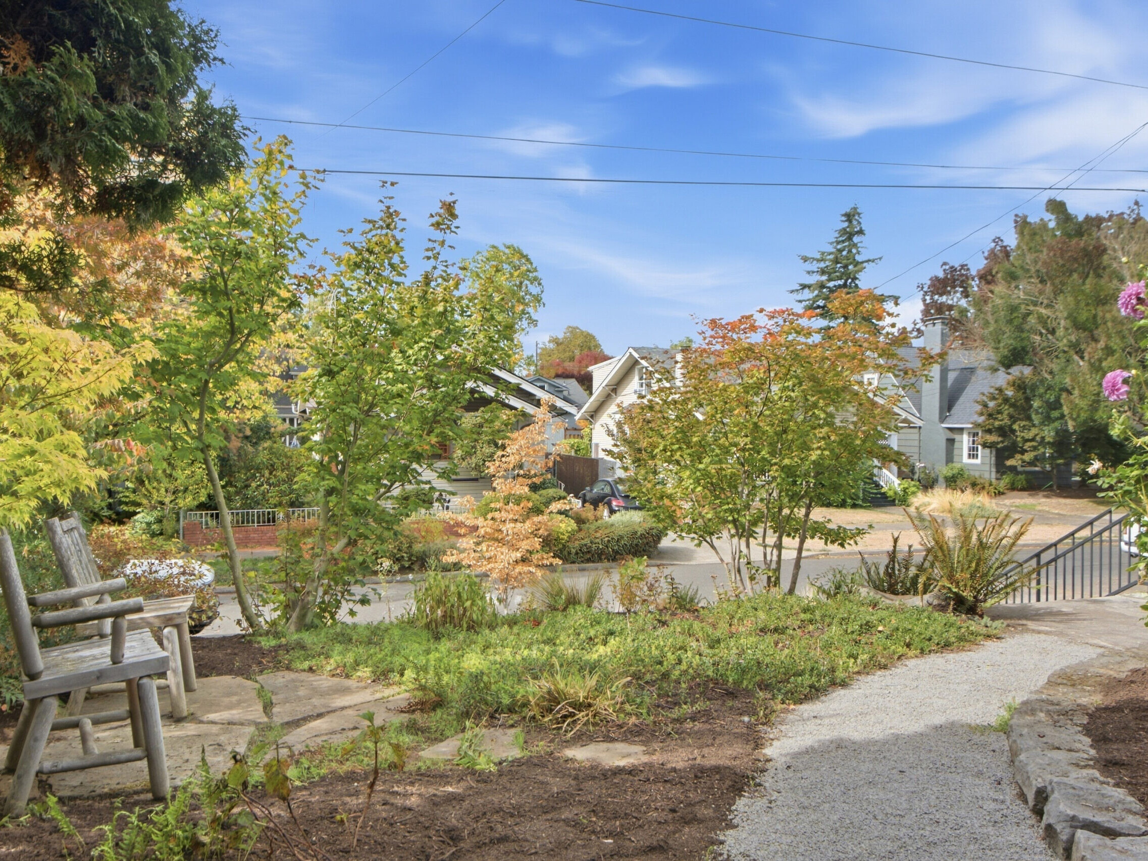 A wooden bench sits in a garden with blooming plants and trees, facing a suburban street lined with houses under a blue sky with wispy clouds. A gravel path curves through the landscaped yard.