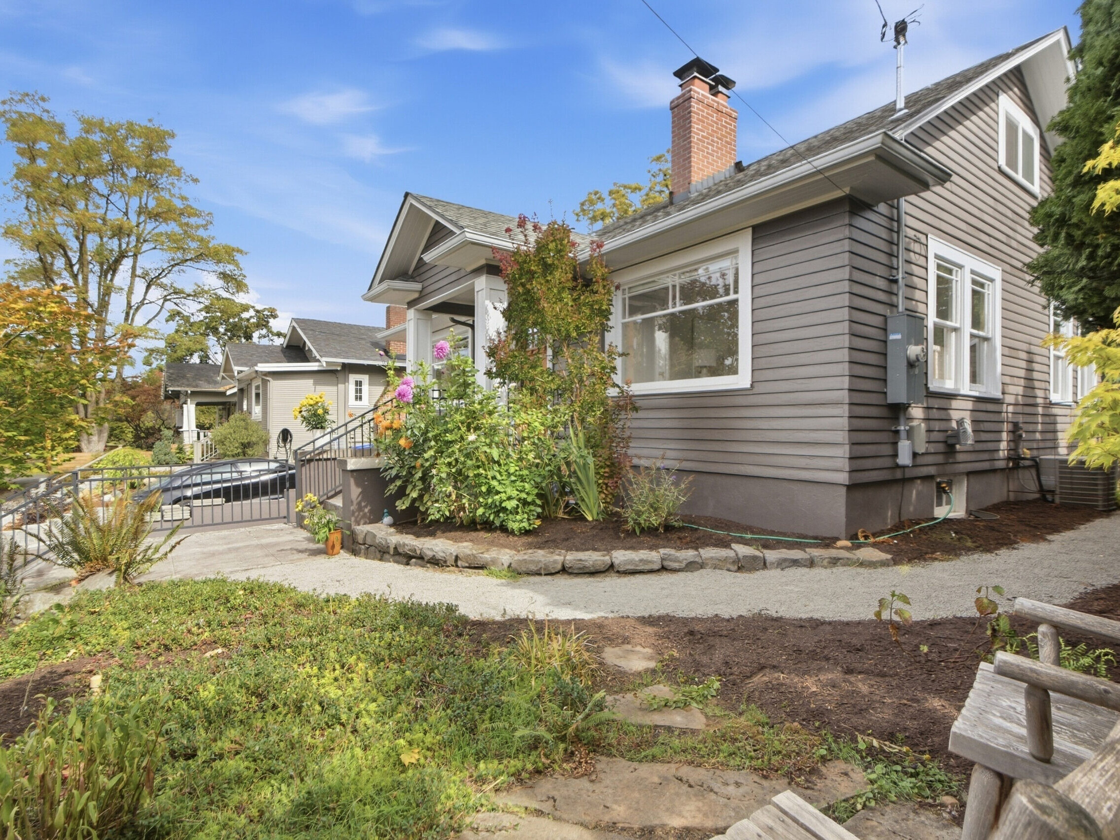 Gray house with white trim, brick chimney, and a landscaped yard with green plants and flowers. A curved walkway leads to the front steps. There is a wooden bench in the foreground and blue sky above.