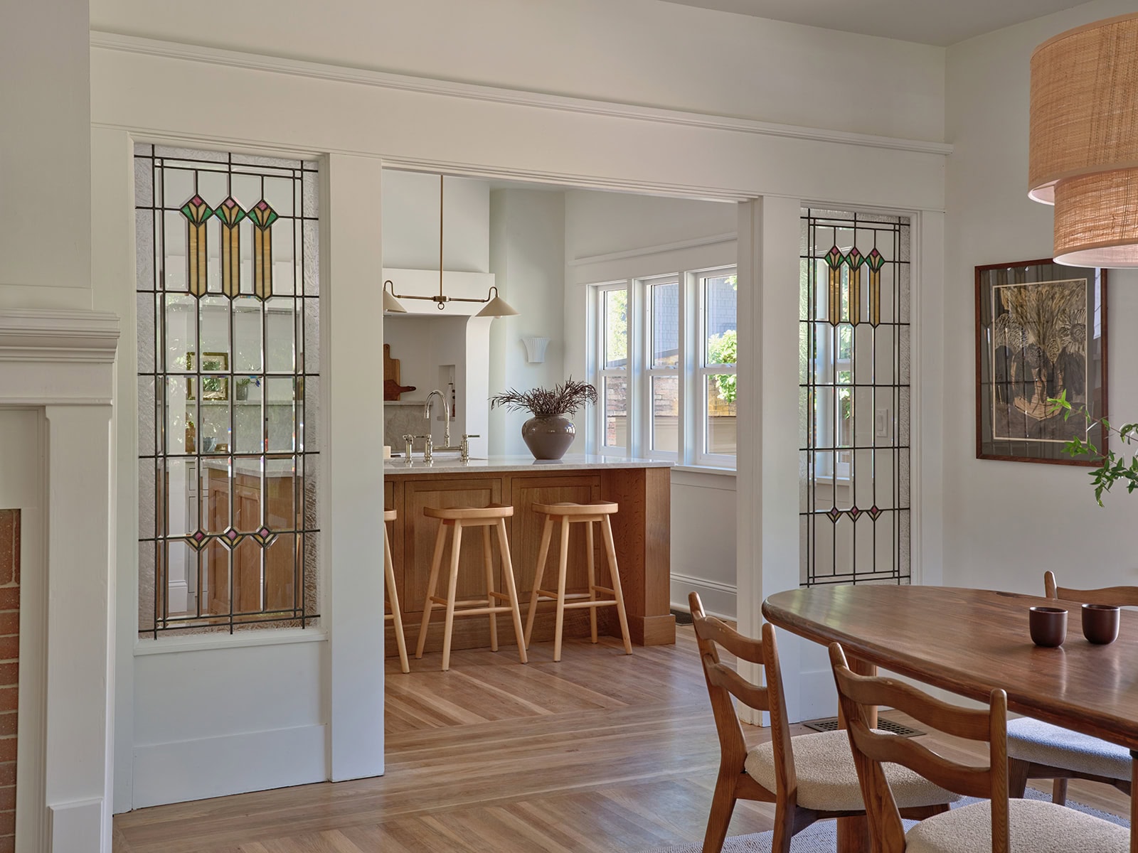 A dining room looking into a kitchen with a wooden island, three stools, and leaded glass windows flanking the doorway.