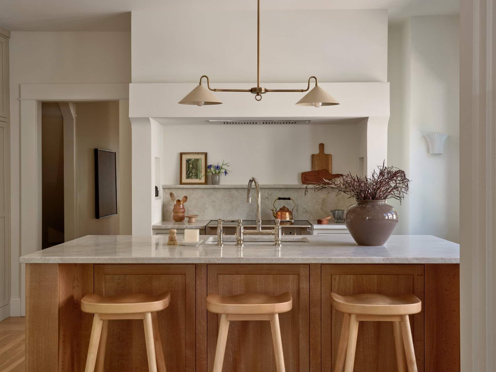 Kitchen island with a marble countertop and three wooden stools, facing a stove recessed into a custom range hood.