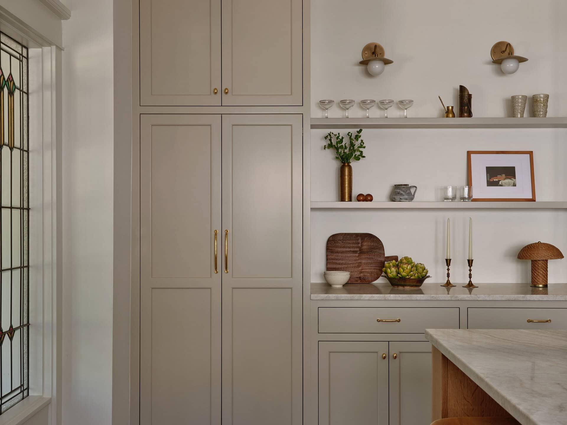Tall beige kitchen cabinets next to white open shelving with glassware, art, and decor, beside a kitchen island with two wooden stools.