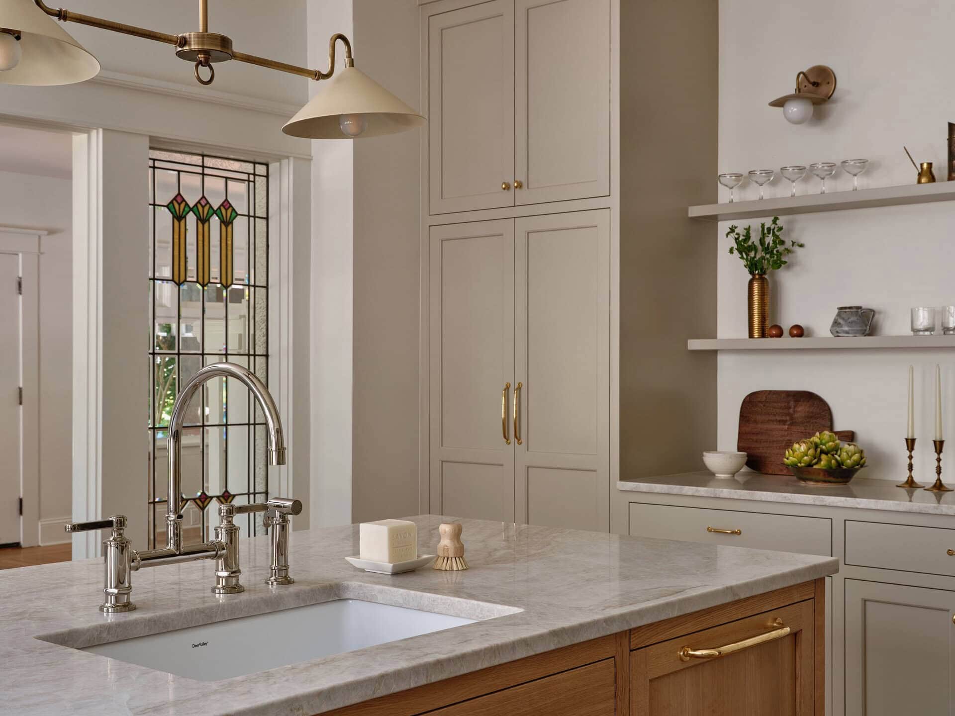 A kitchen island with a marble countertop, undermount sink, and wooden cabinetry, featuring a stained-glass window and open shelving in the background.