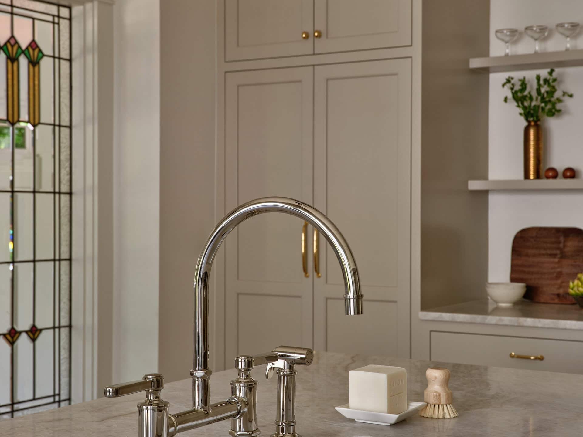 A chrome kitchen faucet with a side sprayer, a bar of soap, and a wooden scrub brush on a stone countertop above a white farmhouse sink.
