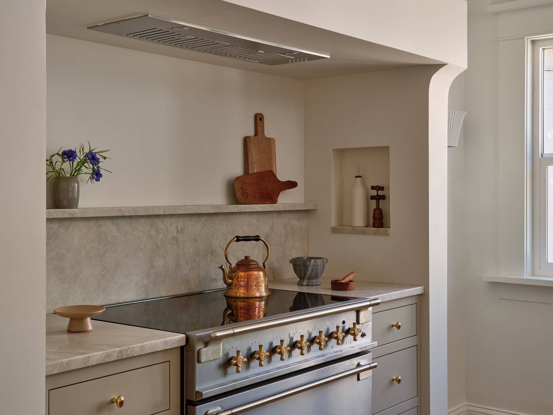 A blue AGA range with a copper tea kettle sits beneath a custom hood in a neutral kitchen with marble counters.