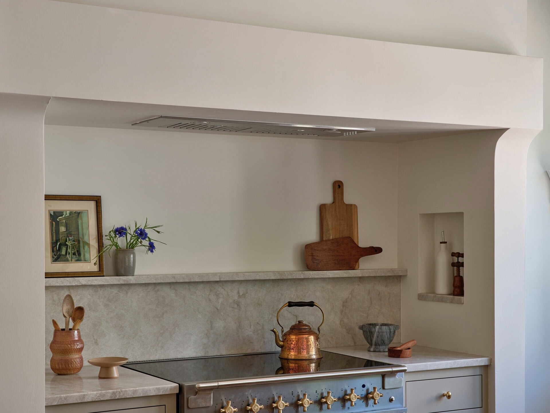 A kitchen stove with a copper kettle, set into a range niche with a stone backsplash, wooden cutting boards, and decorative accessories.
