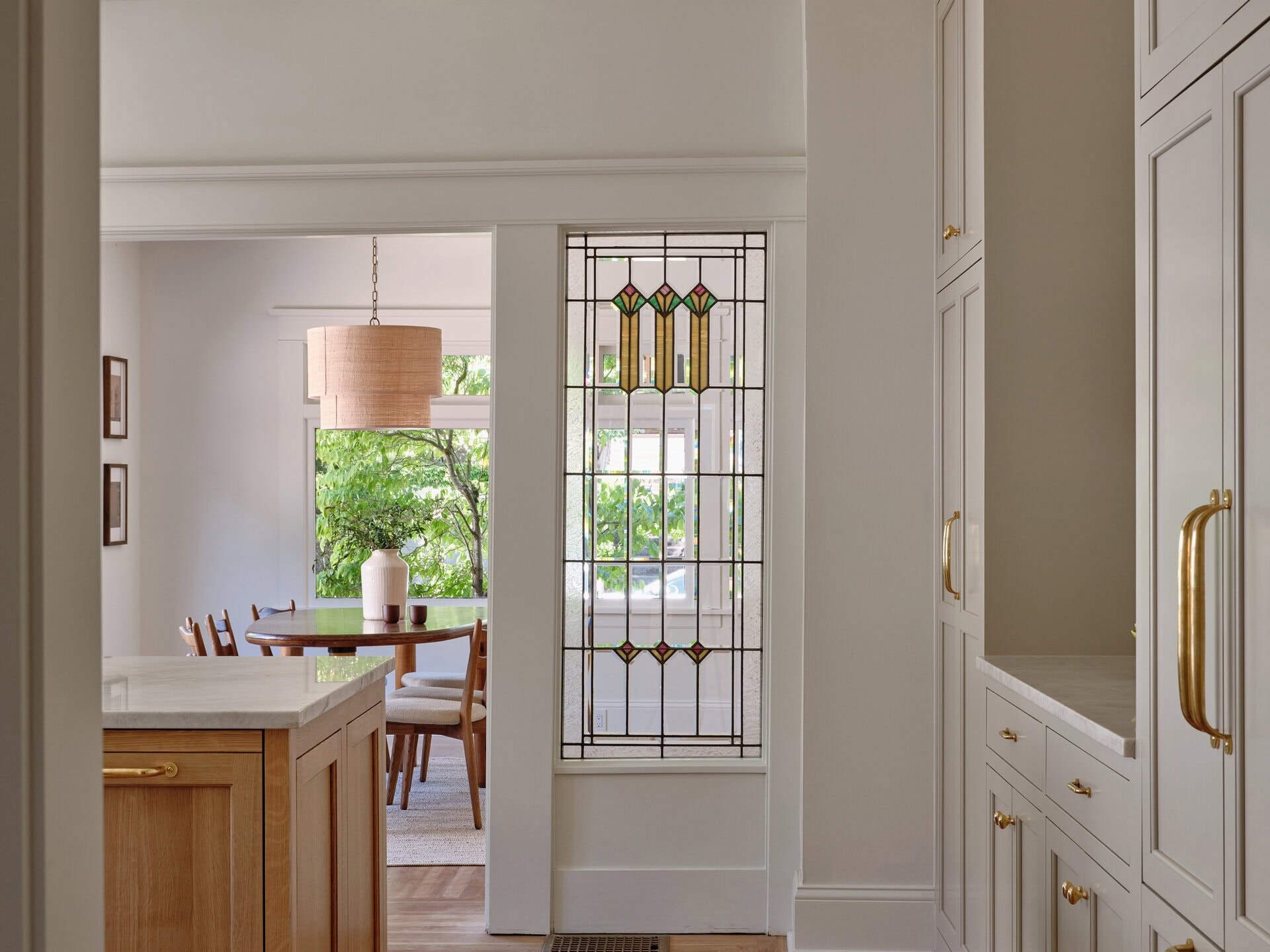 A view from a kitchen with light-colored cabinets and a wooden island into a dining room featuring a round table and a tall stained-glass window panel.