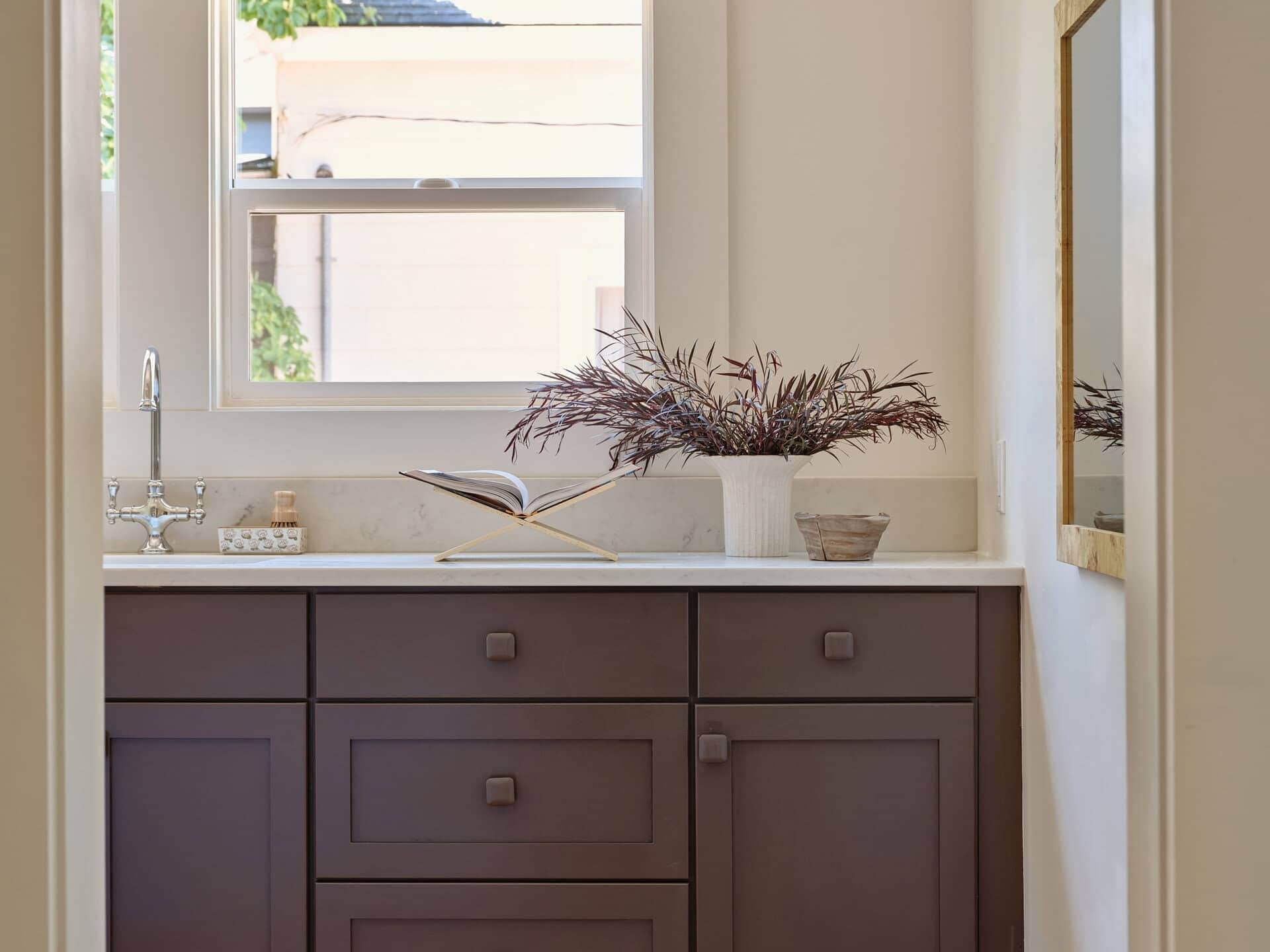A small kitchen area with dark brown cabinets, a white countertop with an open cookbook and vase of foliage, a stainless steel faucet, and a view through a window.