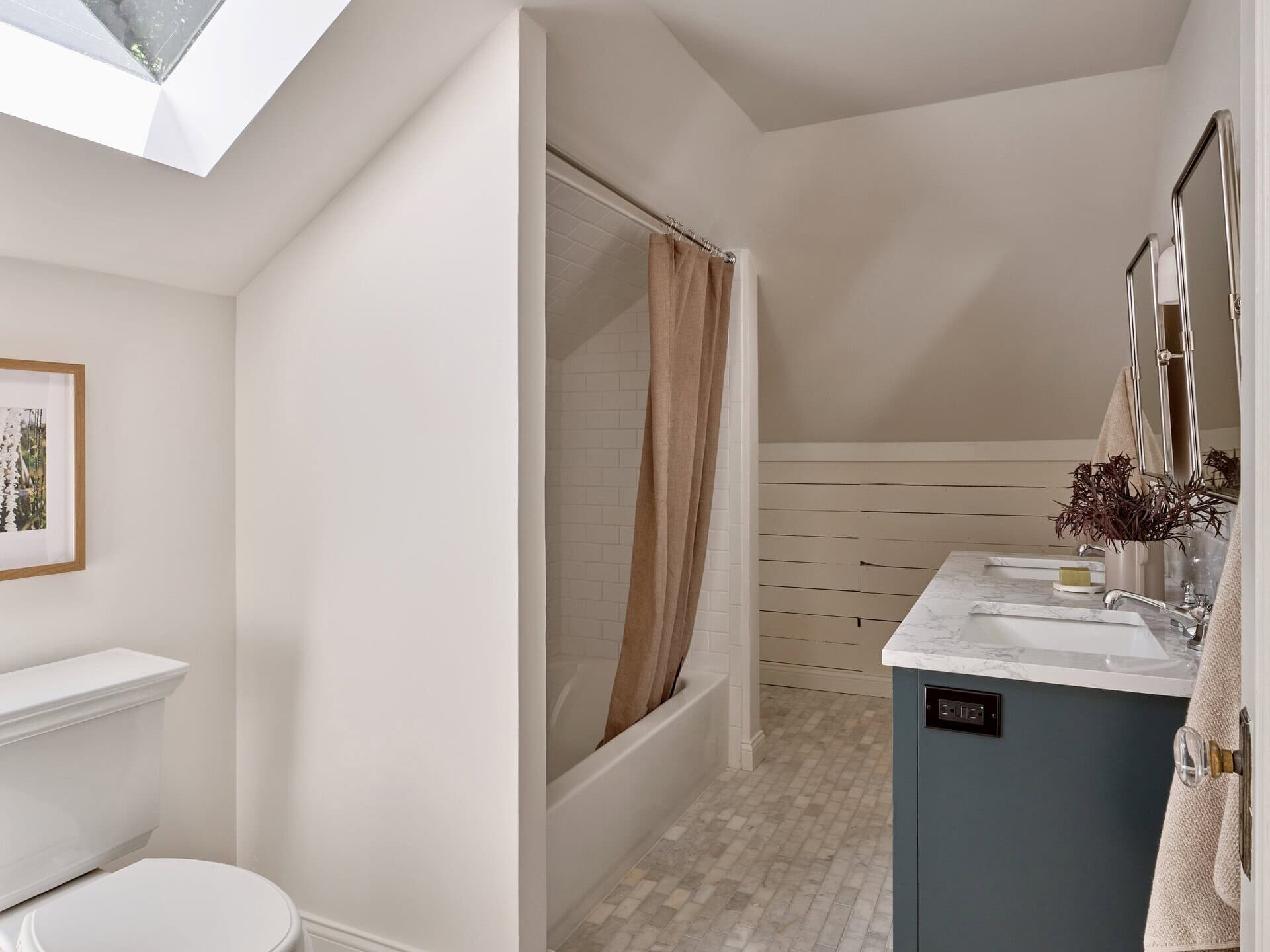 A bathroom with a skylight, a toilet, a bathtub with a curtain, and a dark blue vanity with a marble countertop.