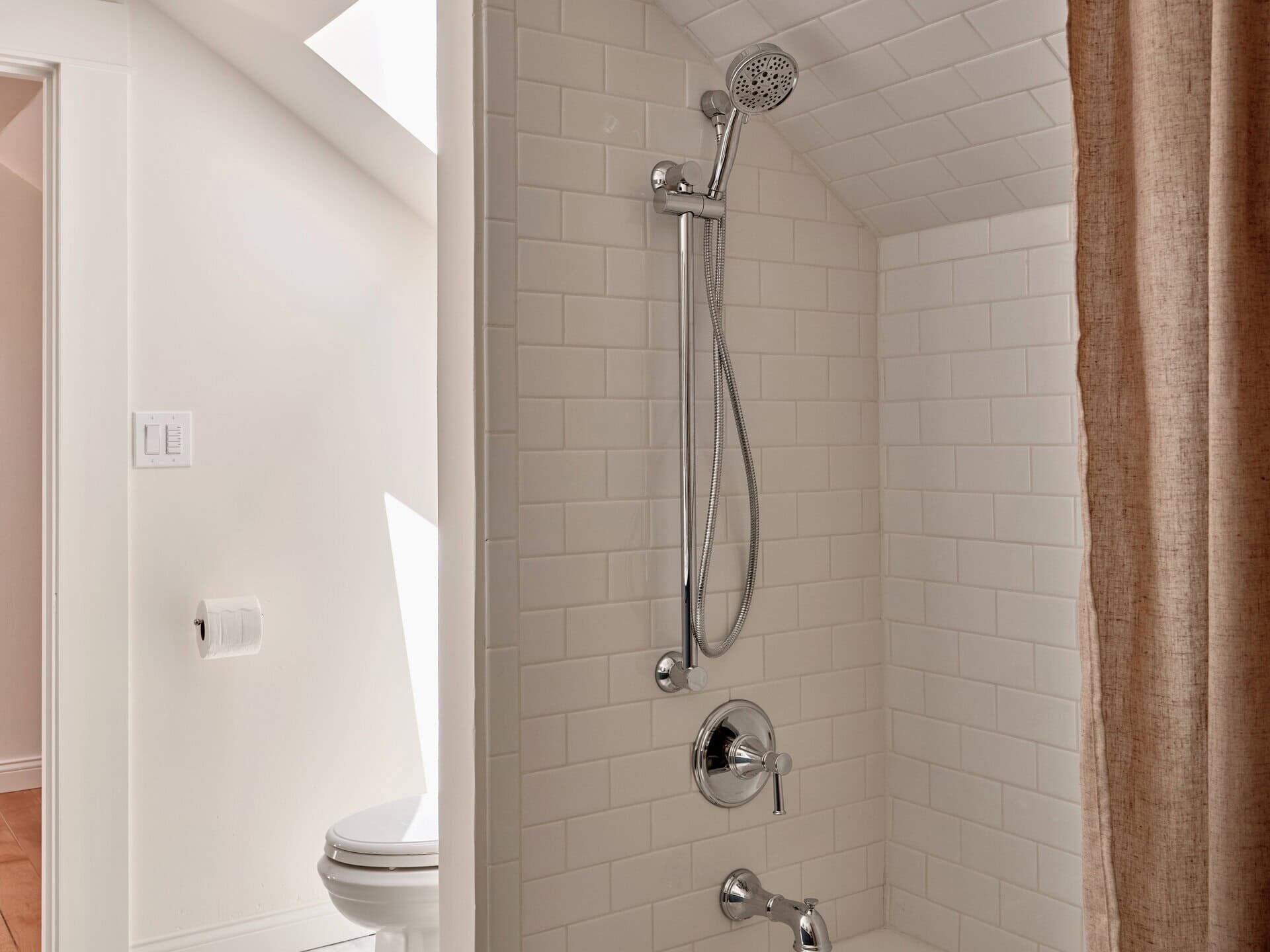A white tiled bathtub with a shower head and a partial curtain, beside a white toilet in a bright bathroom.