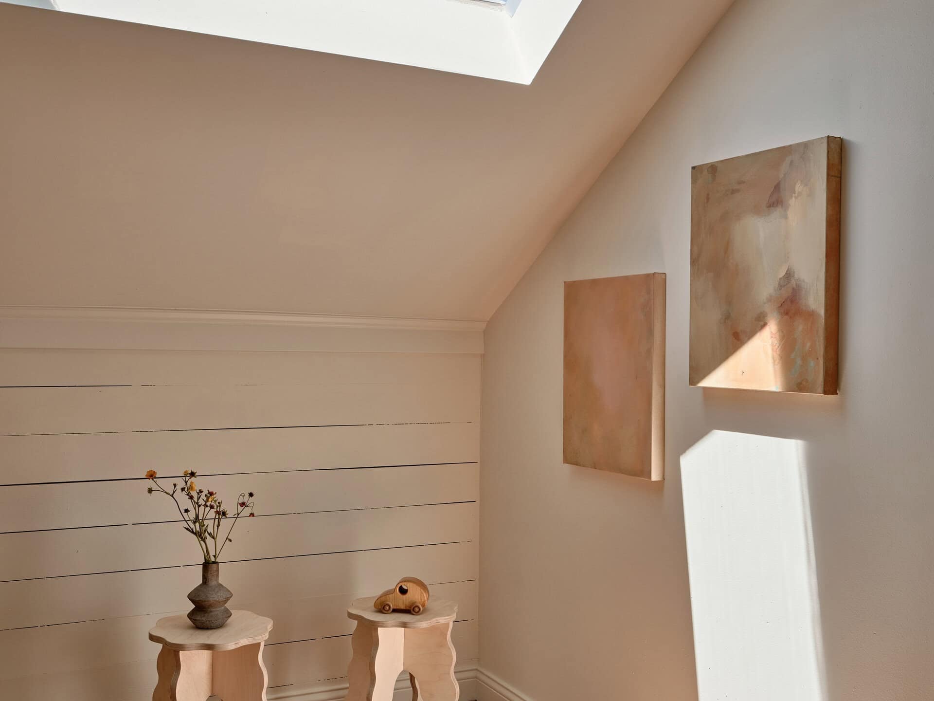 Two wavy-legged wooden stools sit on a hardwood floor beneath a skylight, with a small vase of dried flowers and a wooden toy car on top, and two abstract paintings hanging on the wall.
