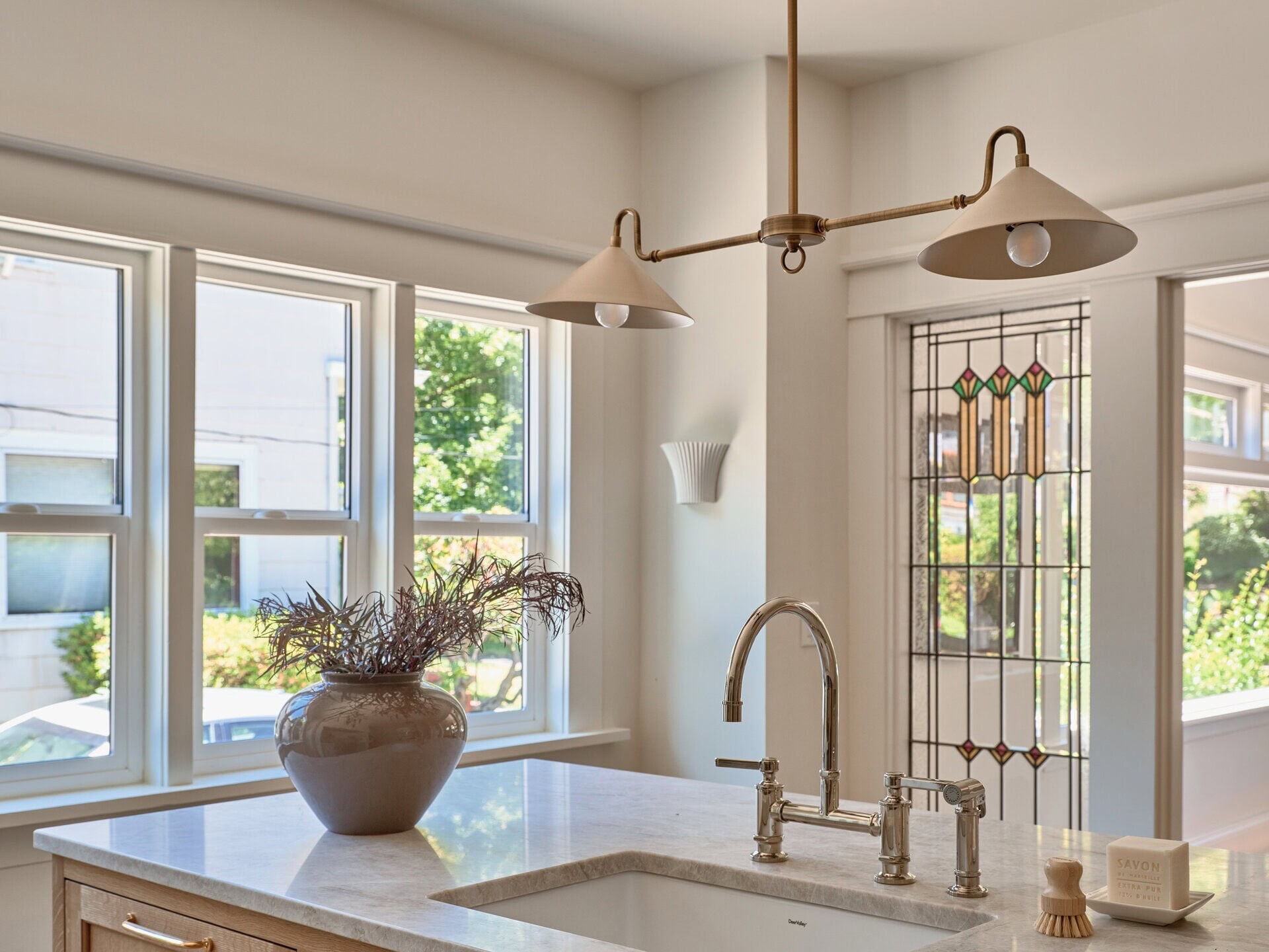 A light wood kitchen island with a marble countertop, undermount sink, and brass faucet, featuring a vase with dried branches and a dish soap block.