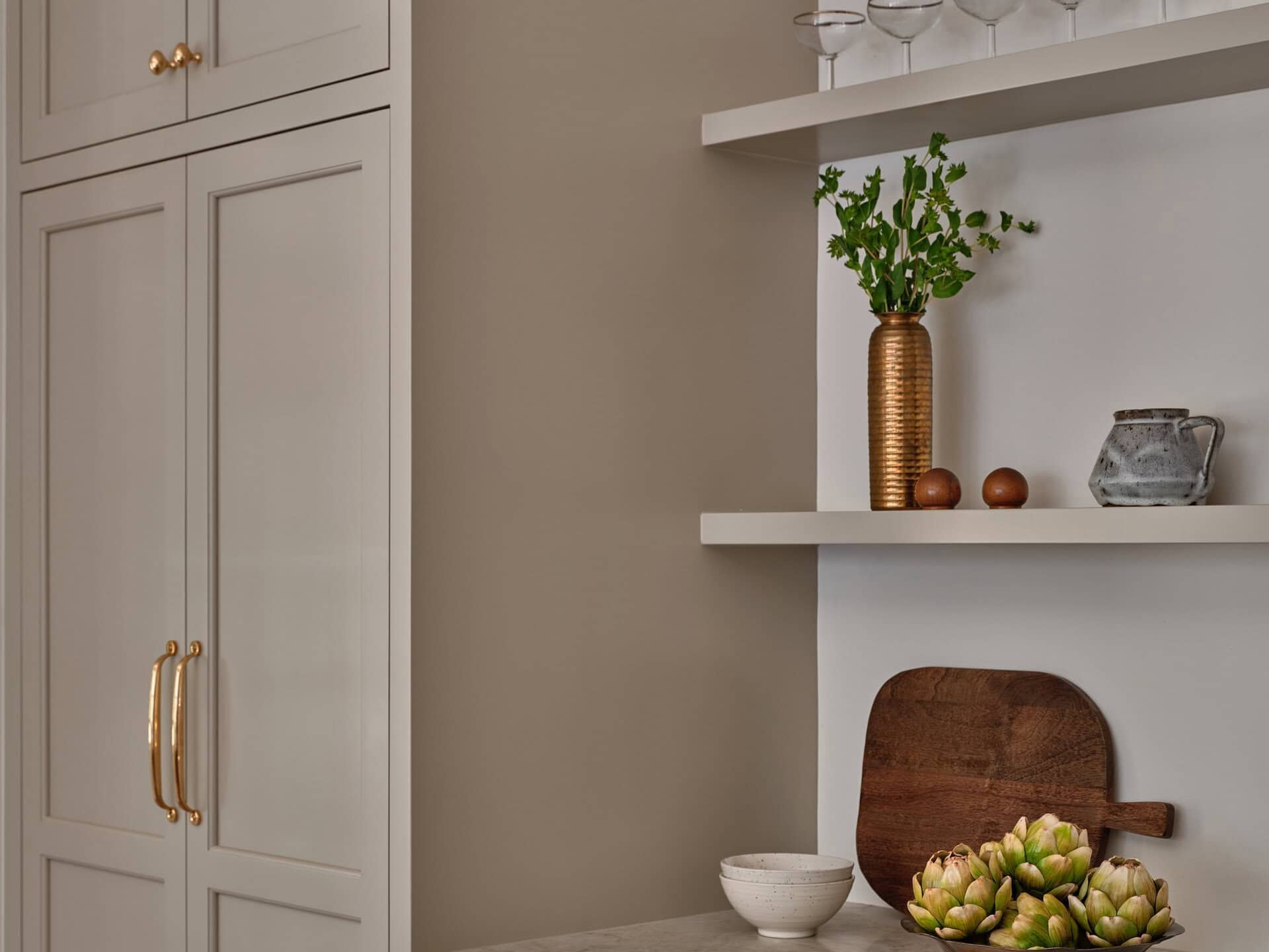 Tall cabinets with gold hardware beside a countertop featuring a bowl of artichokes, a wooden cutting board, a bowl, and open shelving holding glassware and a vase.